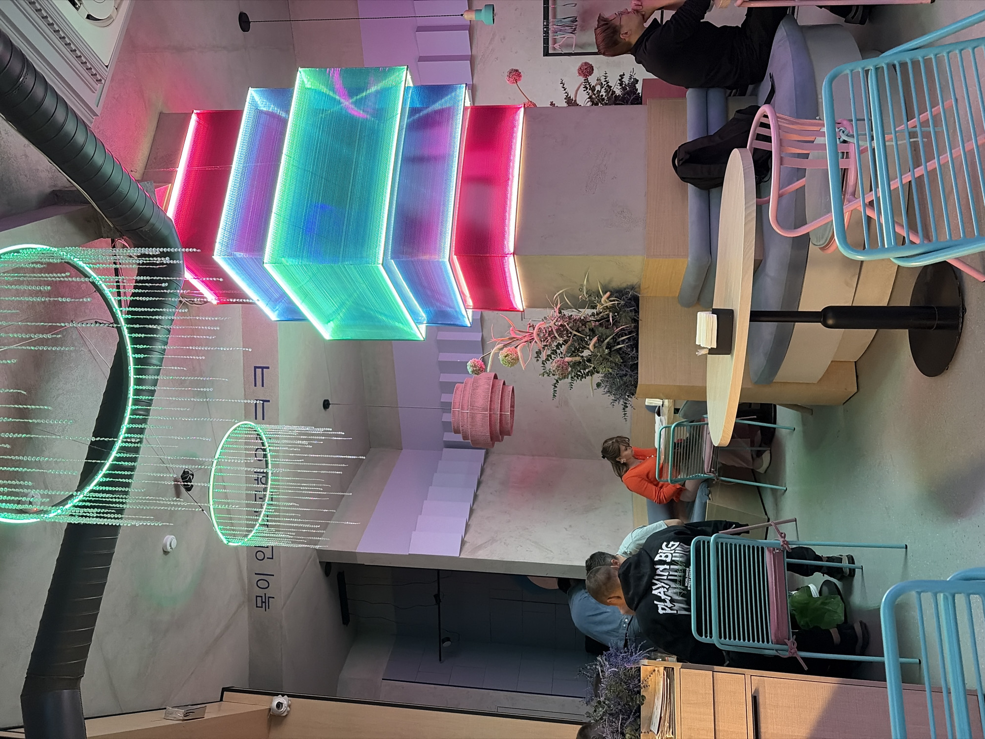 The image captures the interior of a brightly lit, modern cafe in Izhevsk, Russia, characterized by its eclectic and colorful design. In the upper portion of the frame, a concrete ceiling is visible, featuring an exposed dark grey ventilation duct running horizontally. Two large circular neon green light fixtures, adorned with numerous dangling clear beaded strands, hang from the ceiling. A white smoke detector is positioned between these fixtures. On a light grey wall to the left, stylized Korean text is inscribed, reading "목이 인다고 ㅋㅋㅋㅋ." A small white security camera is mounted on the upper left wall. A prominent central concrete pillar extends from the floor, topped with a multi-tiered rectangular light structure. This structure consists of three distinct segments, each illuminated with a different neon color: the top segment is teal, the middle is blue/purple, and the bottom is magenta/red. At the base of this pillar, planters contain arrangements of dried or artificial plants, some with spherical pink elements. The left side of the foreground features a person facing away from the camera, wearing a black hoodie with white text that reads "PLAYIN IN BIG" and additional text below it. This individual is seated in a turquoise slatted metal chair, to which a green plastic bag is tied. They are positioned at a light wooden counter or table. Partially visible behind them is another person wearing a light blue/grey t-shirt. On the right side of the frame, a woman with dark hair is seated on a light blue upholstered bench, wearing an orange long-sleeved top and looking downwards, possibly at a menu or device. A small, round, light wood table with a black base and a black napkin holder is in front of her. Adjacent to this table is a curved pink metal chair. Further to the right, a man wearing a black long-sleeved shirt and glasses is seated on a similar light blue upholstered bench. He rests his right hand on his chin, looking towards the right out of the frame. A black backpack rests on the bench beside him. The right wall features a strip of white and lavender geometric paneling near the top, below which a pink woven pendant lamp hangs. A smaller, light green pendant lamp is visible further right. A clothing rack with hangers is partially visible on the far right edge of the frame. The floor throughout the cafe is a uniform