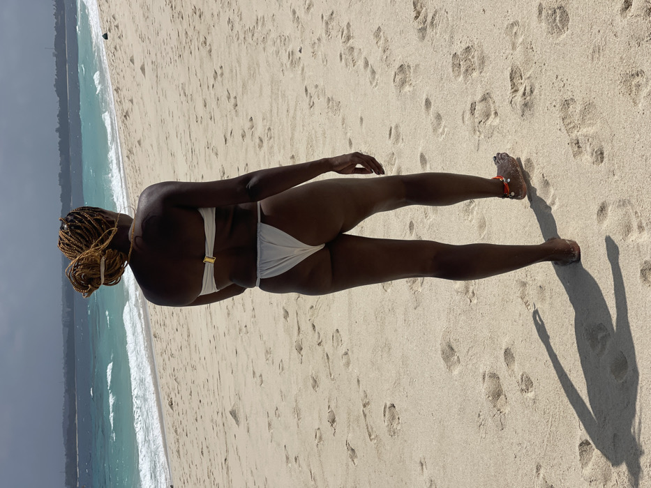 A full-length, rear-view image depicts an individual walking on a sandy beach towards the ocean, located in Zanzibar, Tanzania. The subject has dark skin and light-colored braided hair styled in a bun. They are wearing a light-colored, strapless two-piece bikini; the top features a gold-toned clasp on the back, and the bottom is a high-cut thong style. A large hoop earring is visible on the left ear, and an orange-red string anklet adorned with small white beads is worn on the right ankle. The beach comprises fine, light-colored sand marked with numerous footprints. The ocean in the mid-ground displays clear turquoise and blue water with breaking waves. A distant landmass is discernible on the horizon beneath a partly cloudy sky. Strong overhead sunlight casts a long, distinct shadow of the individual on the sand to their left.