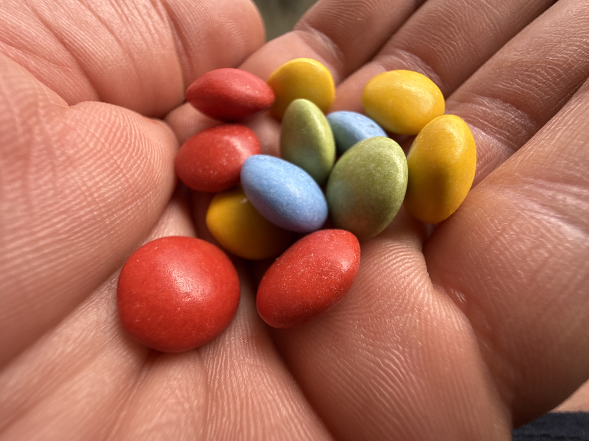 A close-up shot shows a hand holding a collection of colorful candies. The candies are primarily red, yellow, blue, and green. The hand appears to be an adult's hand, with visible skin texture and wrinkles. The environment is not explicitly defined, so no specific location in Strazhitsa, Bulgaria, can be determined from the image alone.
