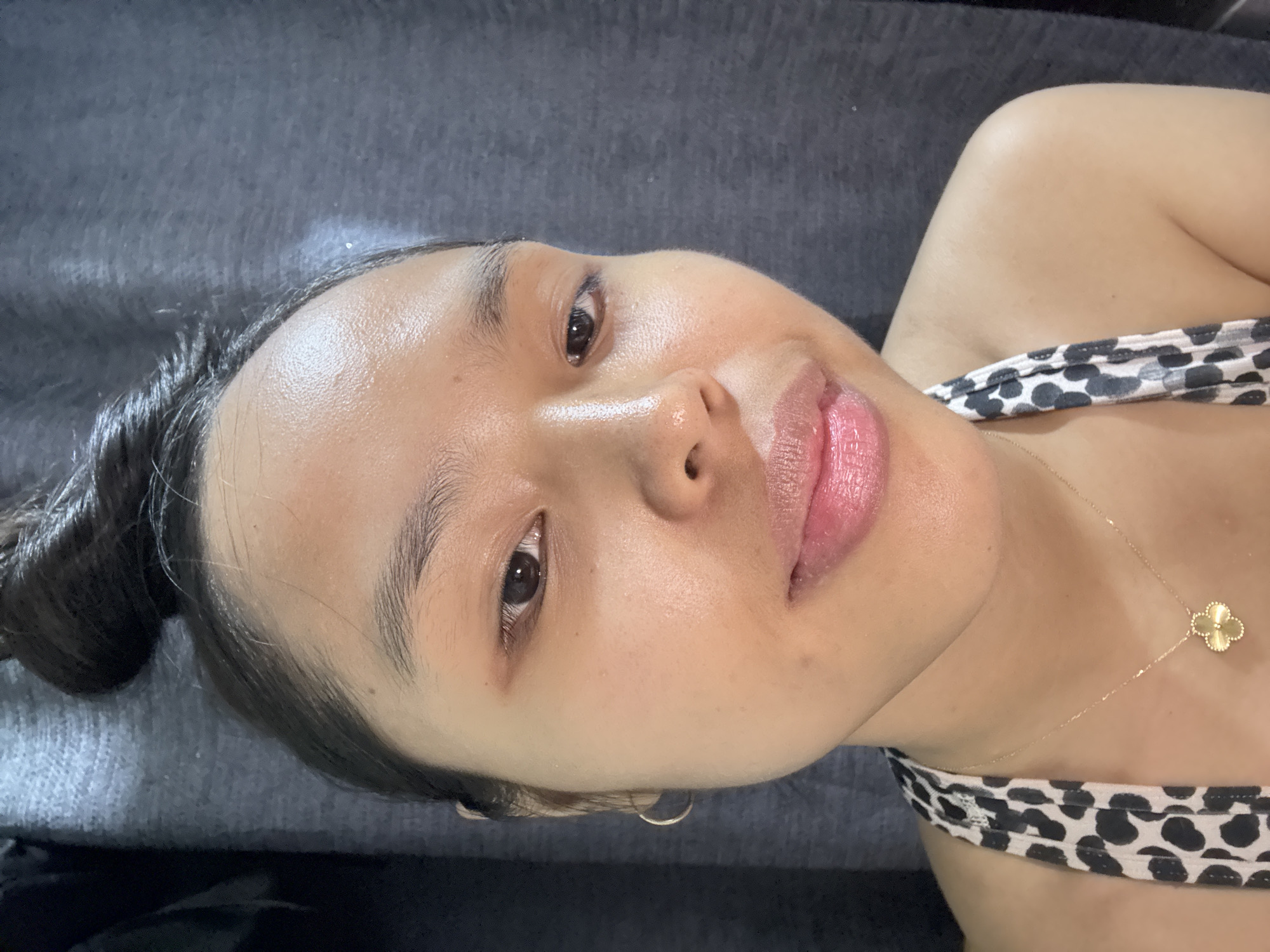 A young woman is lying on a gray surface, likely a bed or sofa. She has dark hair pulled back in a bun, and her skin is glowing. She is wearing a black and white printed top, and a gold necklace with a clover-shaped pendant is visible. She appears to be smiling slightly. There is no information provided in the image to indicate that this photo was taken in Davao City, Philippines.