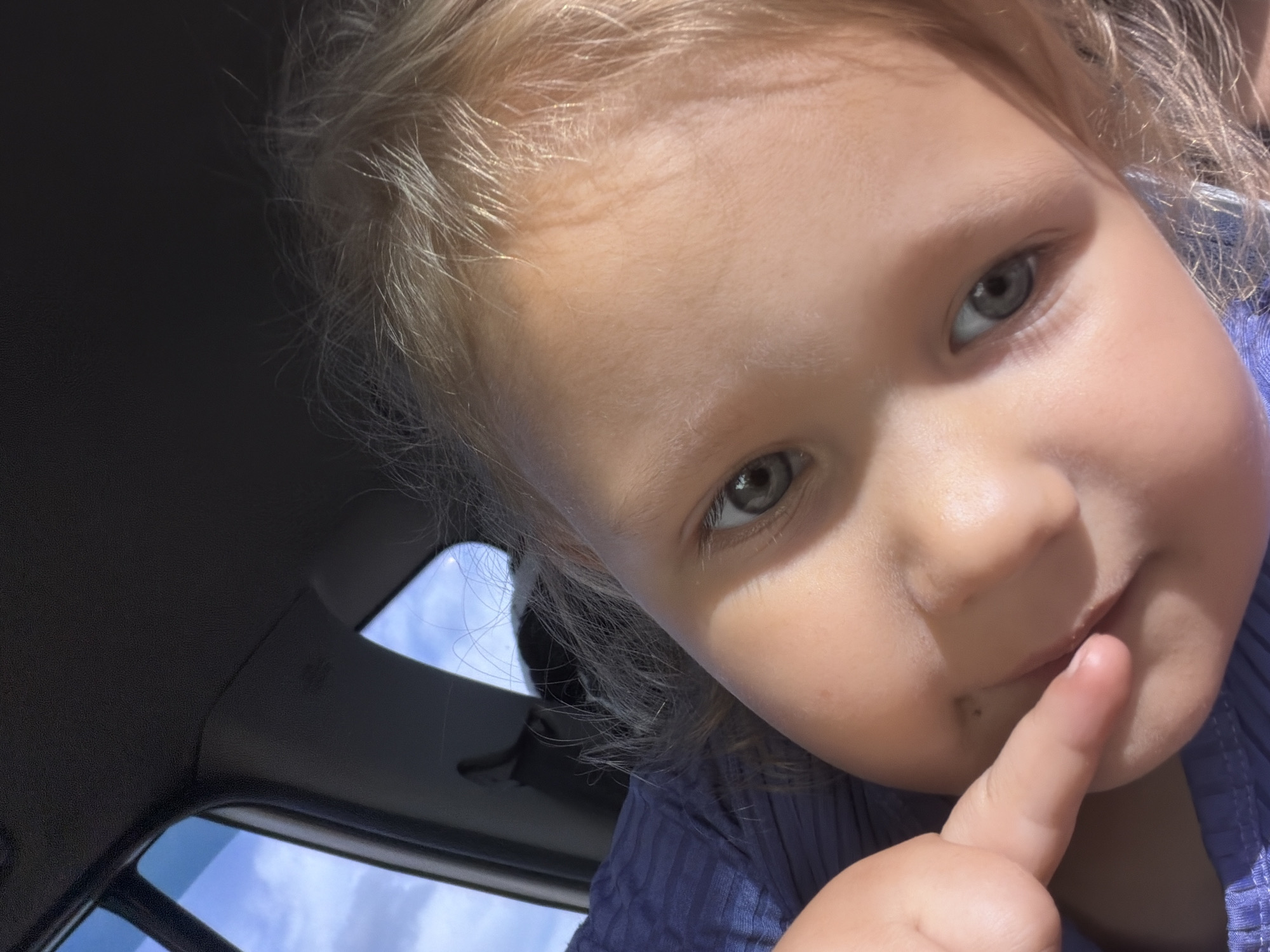 A close-up, low-angle selfie shows a young child with blonde hair looking into the camera. The child's left eye and part of their face are visible, with their finger touching their mouth. The child is wearing a blue shirt. The background appears to be the interior of a vehicle, with the sky and clouds visible through a window. The image quality is fair. The location is not specified in the image or the provided context.