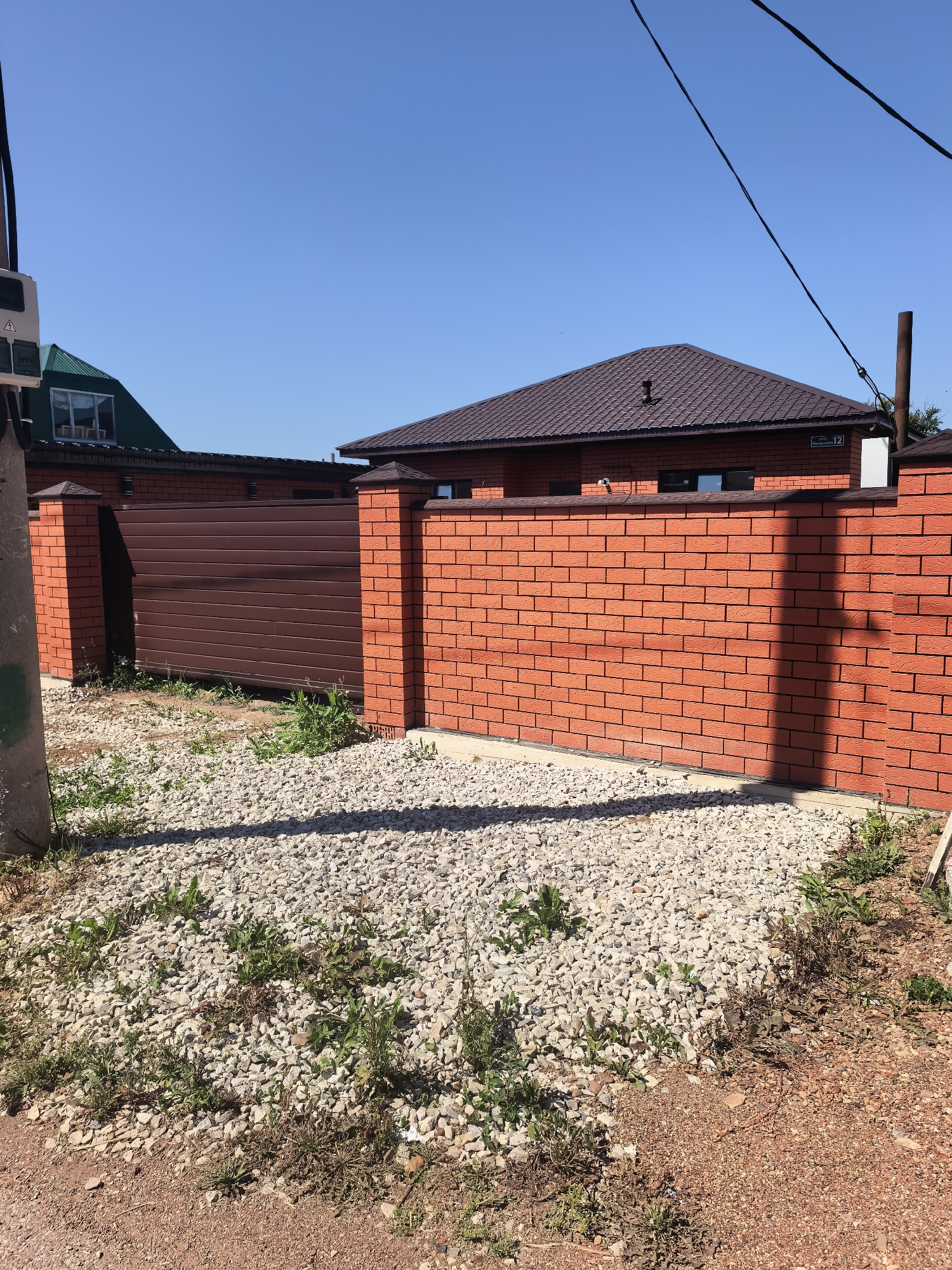 An exterior, eye-level, medium shot captures a sunny day in Sterlitamak, Russia, showcasing a newly constructed brick fence and gate. The fence is made of red bricks with dark mortar, topped with a decorative dark brown capping. To the left, a dark brown metal sliding gate is visible, flanked by brick pillars. Beyond the fence, the roof of a house with dark brown shingles and a chimney is prominent. The ground in front of the fence is covered in light-colored gravel and some sparse green weeds. A utility pole stands to the far left of the frame. The sky is a clear, bright blue, with visible power lines crisscrossing the upper portion of the image. The scene suggests a residential area under development or recently completed construction.