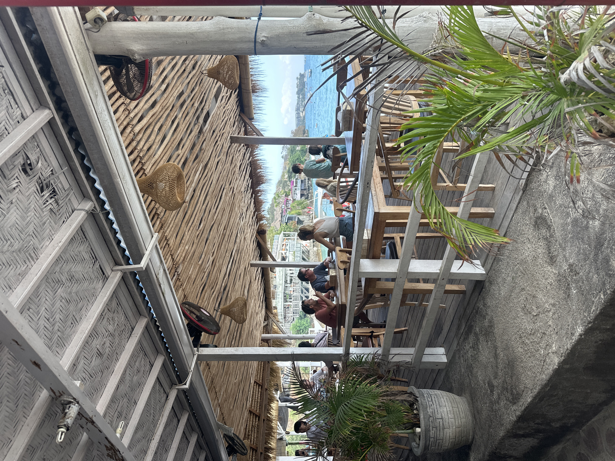 This image depicts an open-air restaurant or beach bar in Nusa Penida, Indonesia, with a thatched roof supported by concrete pillars and wooden beams. The restaurant overlooks a bright blue ocean with several boats visible. In the foreground, a large potted plant with green fronds is situated on a textured concrete surface.
Inside the restaurant, several patrons are seated at wooden tables. On the left, a group of people are dining, with one person wearing a white shirt and glasses, another in a dark shirt, and a third in a light-colored t-shirt. Towards the center, a couple is engaged in conversation at a table, with the male wearing a reddish-orange t-shirt and the female in a grey t-shirt. Further back, a person in a blue shirt is seated at a table. In the background, more diners are visible, and the coastline of Nusa Penida is dotted with buildings.
The restaurant interior features rustic wooden furniture and decorative wicker pendant lights hanging from the ceiling. Industrial-style fans are mounted on the ceiling structure. The flooring appears to be wooden planks. The overall atmosphere suggests a relaxed, tropical dining experience with a view of the sea.