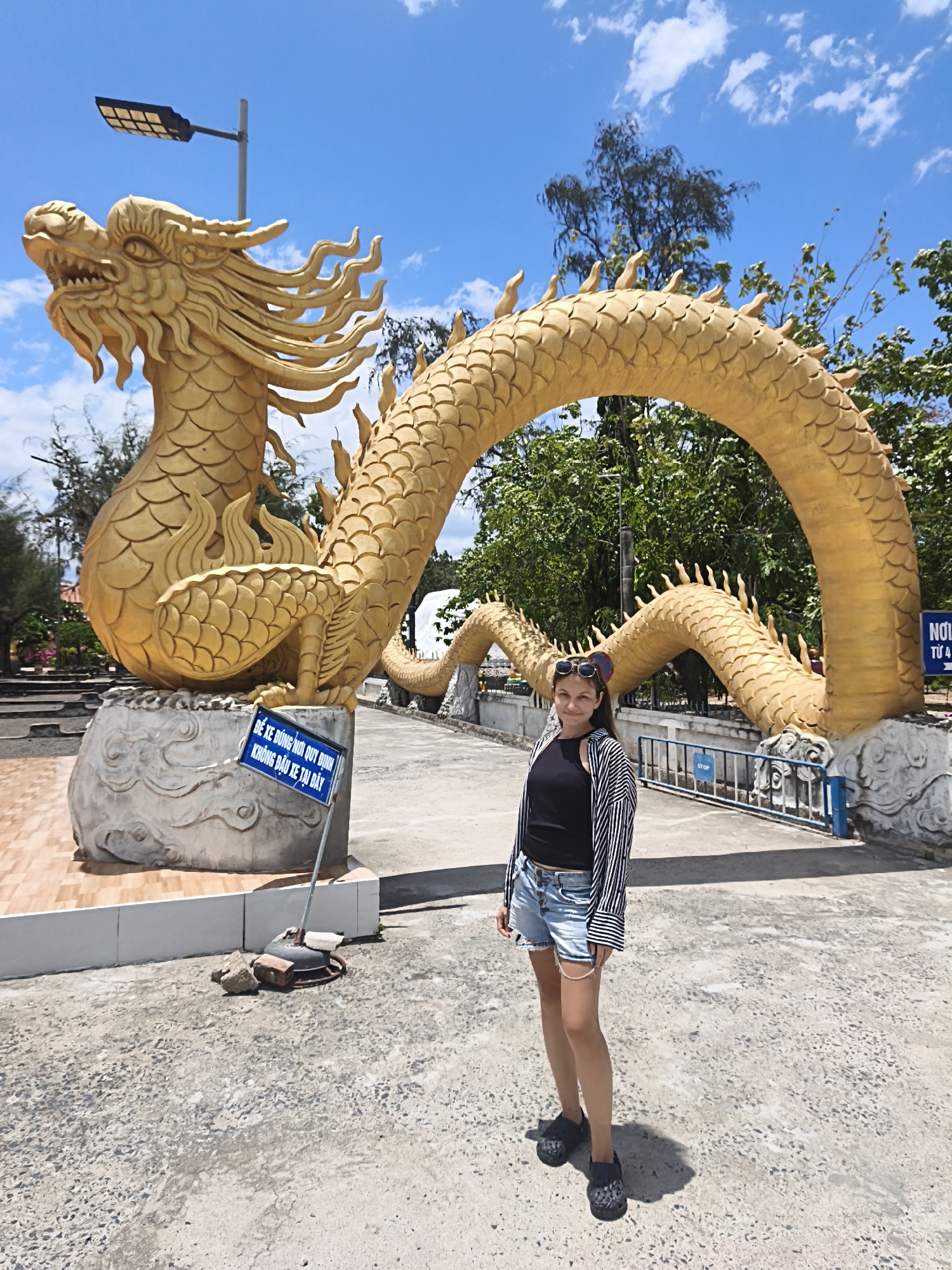 A medium shot captures a young woman standing in front of a large, golden dragon sculpture in Cam Ranh, Vietnam. The dragon is coiled, with its head on the left and its body arching over an open pathway. The woman, wearing a black tank top, a striped open shirt, denim shorts, and dark clogs, stands facing the camera. Her hair is pulled back, and sunglasses are perched on top of her head. A blue sign with Vietnamese text is visible near the dragon's base, and a fence with a "STOP" sign is in the background. The scene is set outdoors with a bright blue sky and scattered clouds overhead, along with trees and foliage in the background. The ground is paved with concrete.