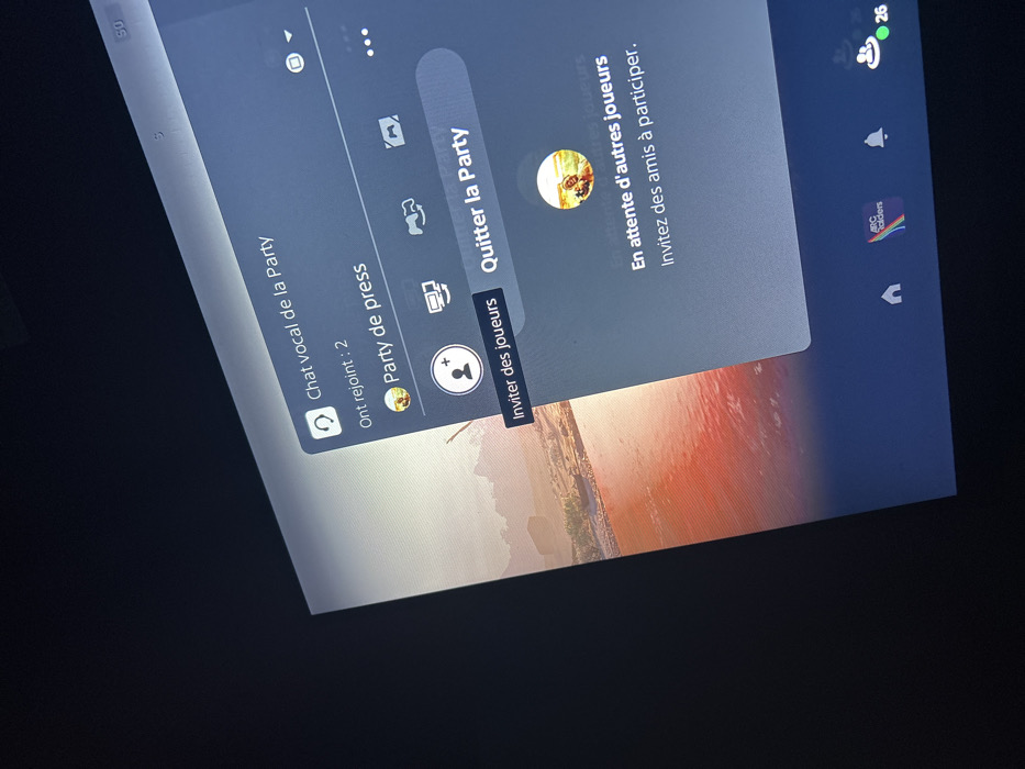 This image captures a digital screen displaying a gaming console interface, likely a PlayStation, within an indoor environment in Illkirch-Graffenstaden, France. The screen is angled, showing a semi-transparent dark blue overlay menu on the right. This menu, titled "Chat vocal de la Party" (Party voice chat), indicates "Ont rejoint : 2" (Joined: 2) and specifies the group as "Party de press" (Press Party). Visible interactive elements include buttons labeled "Inviter des joueurs" (Invite players) and "Quitter la Party" (Leave Party). A message states, "En attente d'autres joueurs Invite des amis à participer." (Waiting for other players Invite friends to participate.). A circular profile icon depicts a figure on an animal within a landscape scene. The background behind the menu shows a digital landscape featuring reddish-orange water, rock formations, and what appears to be a desert environment. The bottom edge of the screen displays a row of interface icons: a home symbol, an application icon with "ABC builders" and a rainbow logo, a notification bell, and a user icon showing a green status dot and the number "26". The surrounding area outside the screen is dark, indicating a dim room.