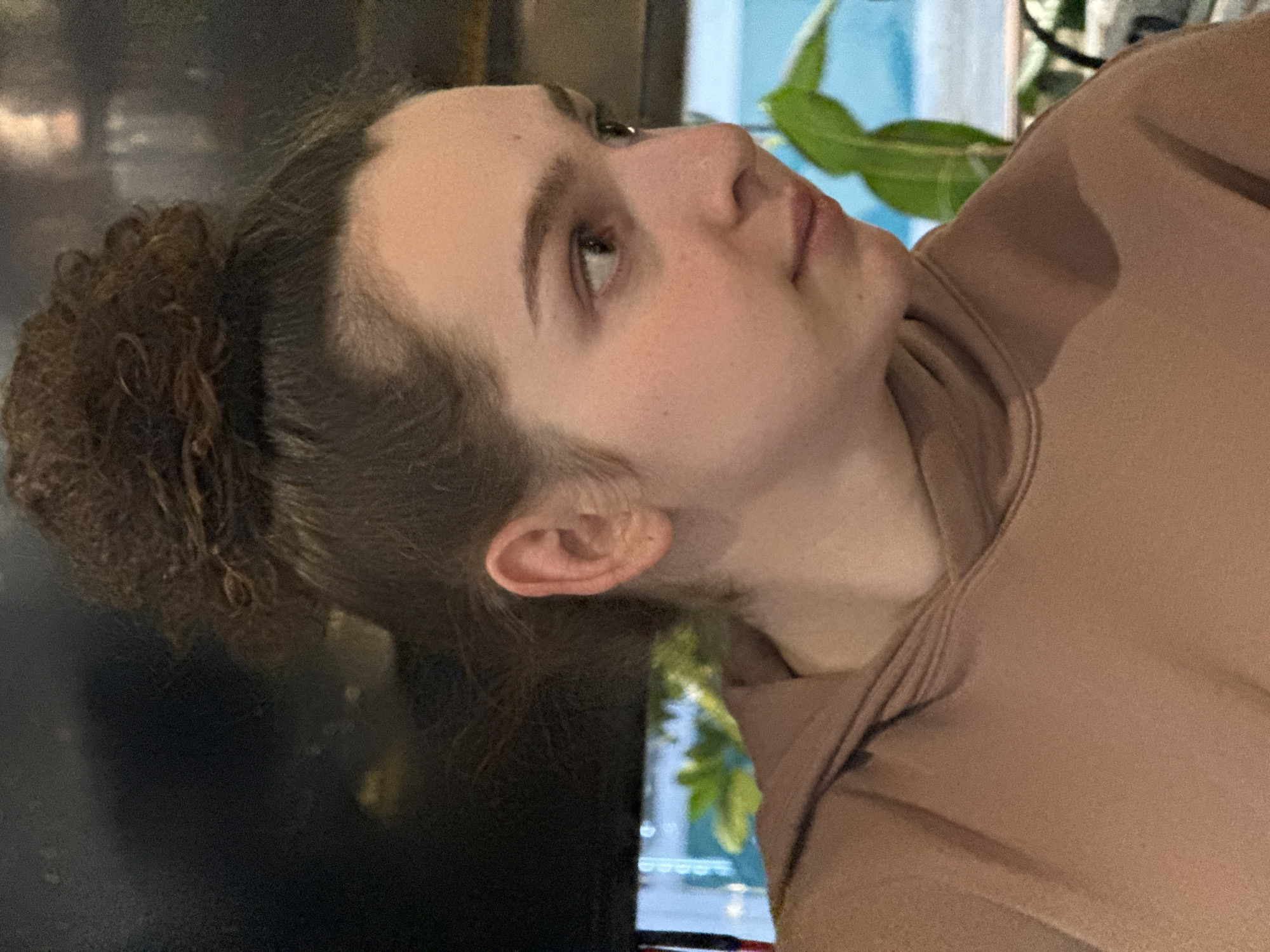 The image displays a young female in right profile, from the upper chest to the top of her head, situated indoors in Sofia, Bulgaria. Her dark brown hair is gathered into a high, messy bun. She possesses fair skin with visible freckling on her right cheek and light brown eyes, which are directed slightly downward and to the right. Her lips are closed. She is attired in a plain, light brown, high-necked, long-sleeved garment, consistent with a hoodie or sweatshirt. The background to her right consists of out-of-focus green foliage and a bright blue section, indicative of an outdoor view or an interior element like a large display. The background to her left is dark and indistinct.