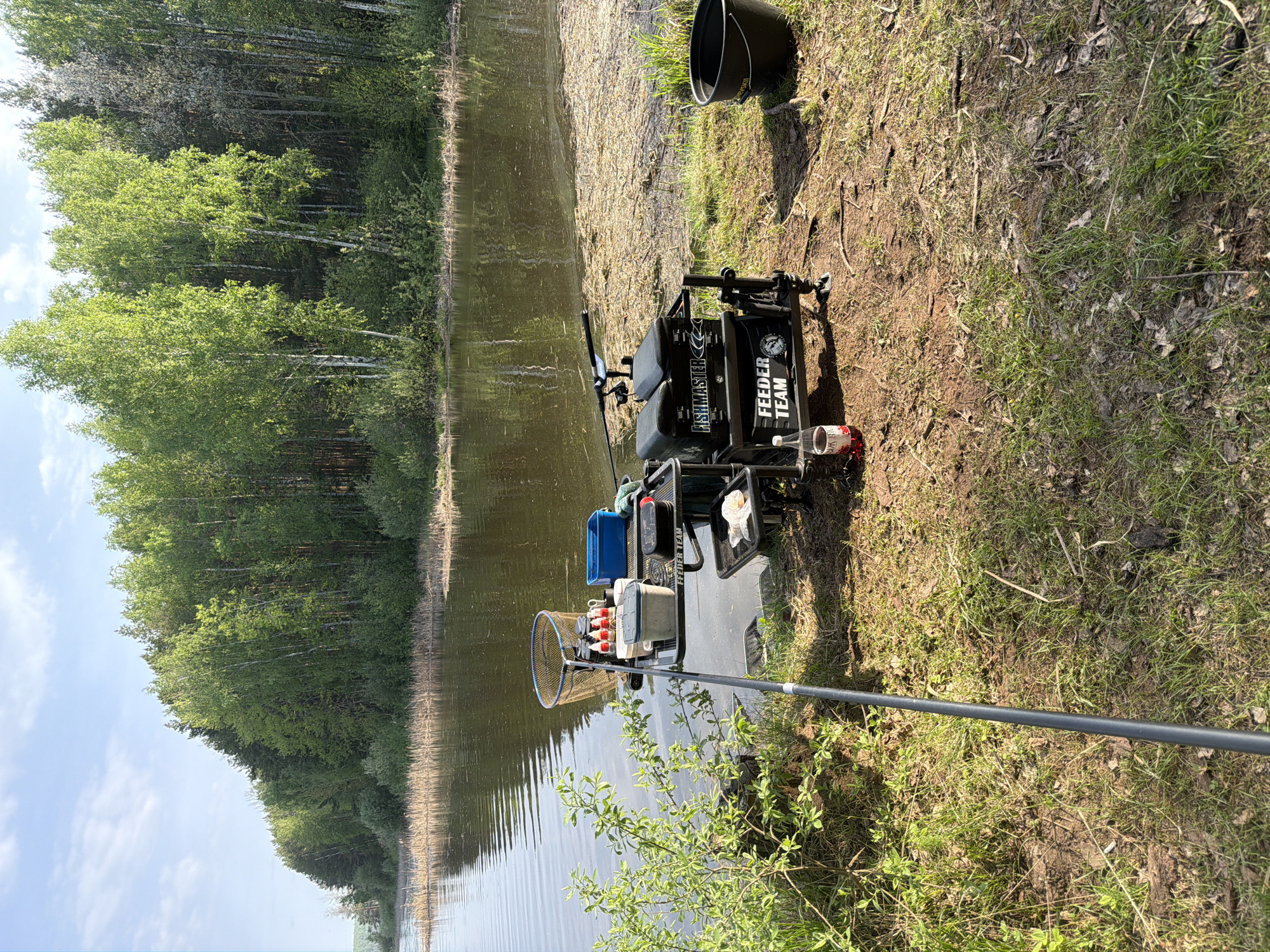 The image shows a fishing setup at a lake. The location is likely in a rural area, given the dense forest in the background. A body of water is in the foreground, with trees reflected in its surface.
On the bank of the lake, a fishing station is set up. The fishing station is black and features the text "FISHMASTER" and "FEEDER TEAM". A black bucket rests next to the fishing station. A long fishing rod extends towards the lake, while a landing net is positioned near the edge of the water. A blue container, a white container, and several red containers are on the fishing station.