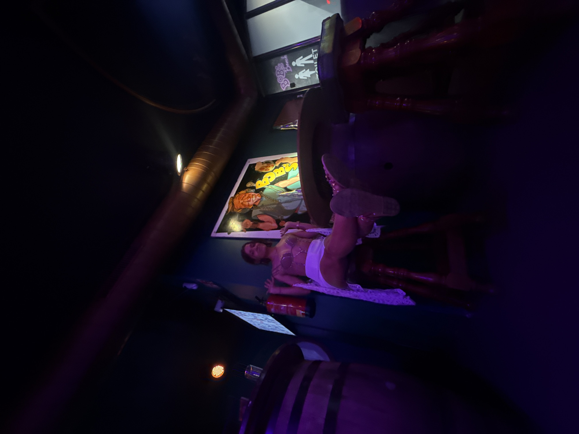 This image captures an indoor scene at a bar or club, identified as located in Calella, Spain. The environment is dimly lit with predominant purple and reddish ambient lighting.
A single adult female is seated on a dark wooden high stool in the center-right of the frame, facing slightly left. She is wearing a light-colored, possibly white, strappy bralette-style top with intricate chain-like detailing across the front, white shorts, and a long, white, lace-patterned garment draped over her legs and extending to the floor. Her bare feet, adorned with metallic-studded sandals, are propped up on the surface of a large, dark wooden barrel that functions as a table. Her arms are raised, with her hands holding the straps of her top near her shoulders, and she displays a slight smile.
To the left of the woman, mounted on a dark teal-colored wall, is a red fire extinguisher. Further left, an illuminated orange-red circular fixture is visible on the wall, and a smaller screen or sign with white text is mounted above it. On the far left, another large wooden barrel, identical in style, serves as a table with a clear glass resting on its surface.
Behind the woman, on the teal wall, a large framed artwork features cartoonish figures, with a prominent orange-haired, bearded male character wearing a light blue garment. To the right of this artwork, a vertical black sign with white text and pictograms reads "Bob's Beach TOILET" accompanied by male and female restroom symbols.
The upper portion of the image reveals large, bronze-colored ventilation ducts or pipes running horizontally across the dark ceiling, with one pipe curving upwards on the right. A focused white spotlight emanates from the ceiling, illuminating the top of the left barrel. The floor is dark and largely obscured.