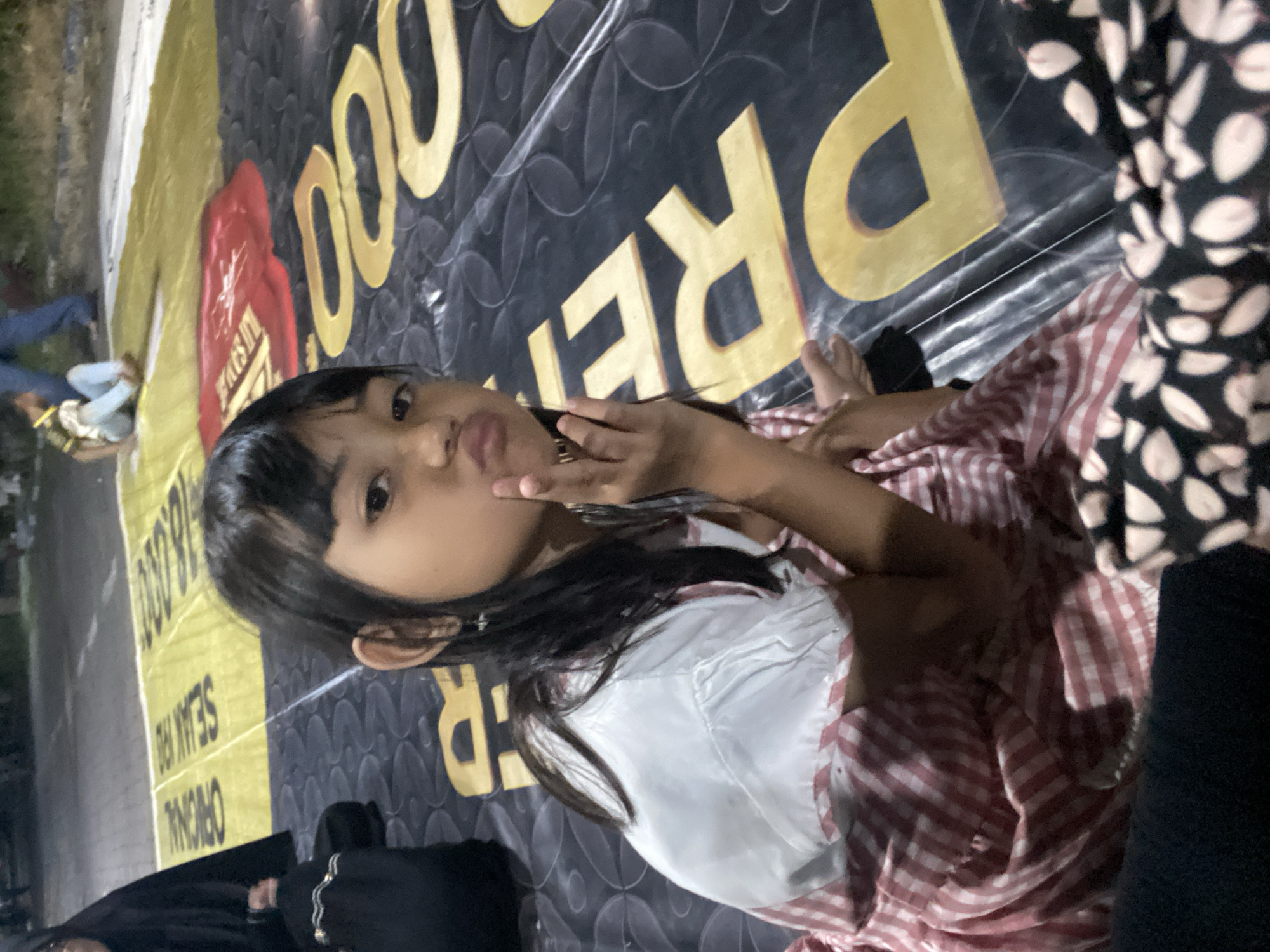 A young girl with dark hair and a pink and white checkered dress poses in front of a backdrop advertising the "ORIGINAL SEAN RO 16.000 DODOPI REJER". The girl is looking at the camera with her lips pursed and her hand slightly raised. Other people are present, but only partially visible on the sides of the frame. The location is identified as Gelam, Indonesia based on the lettering present in the background. The environment appears to be indoors, potentially at a promotional event.