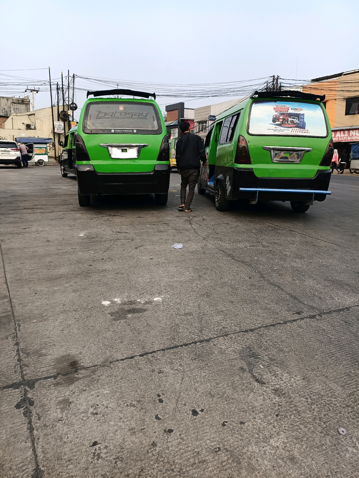 In Tangerang, Indonesia, two green minibuses are parked on a concrete road in front of buildings. The minibus on the left has a spoiler and a sticker on its rear window that says "HAI ROSAY PANCA TUNGGAL". The minibus on the right has a sticker on its rear window featuring two red and white vehicles. A person wearing a black jacket and brown pants walks away from the viewer between the two minibuses. Another person in a pink headscarf and black pants walks away from the viewer on the right side of the image, in front of a white vehicle. The sky is overcast, and there are electrical wires and poles visible in the background.
