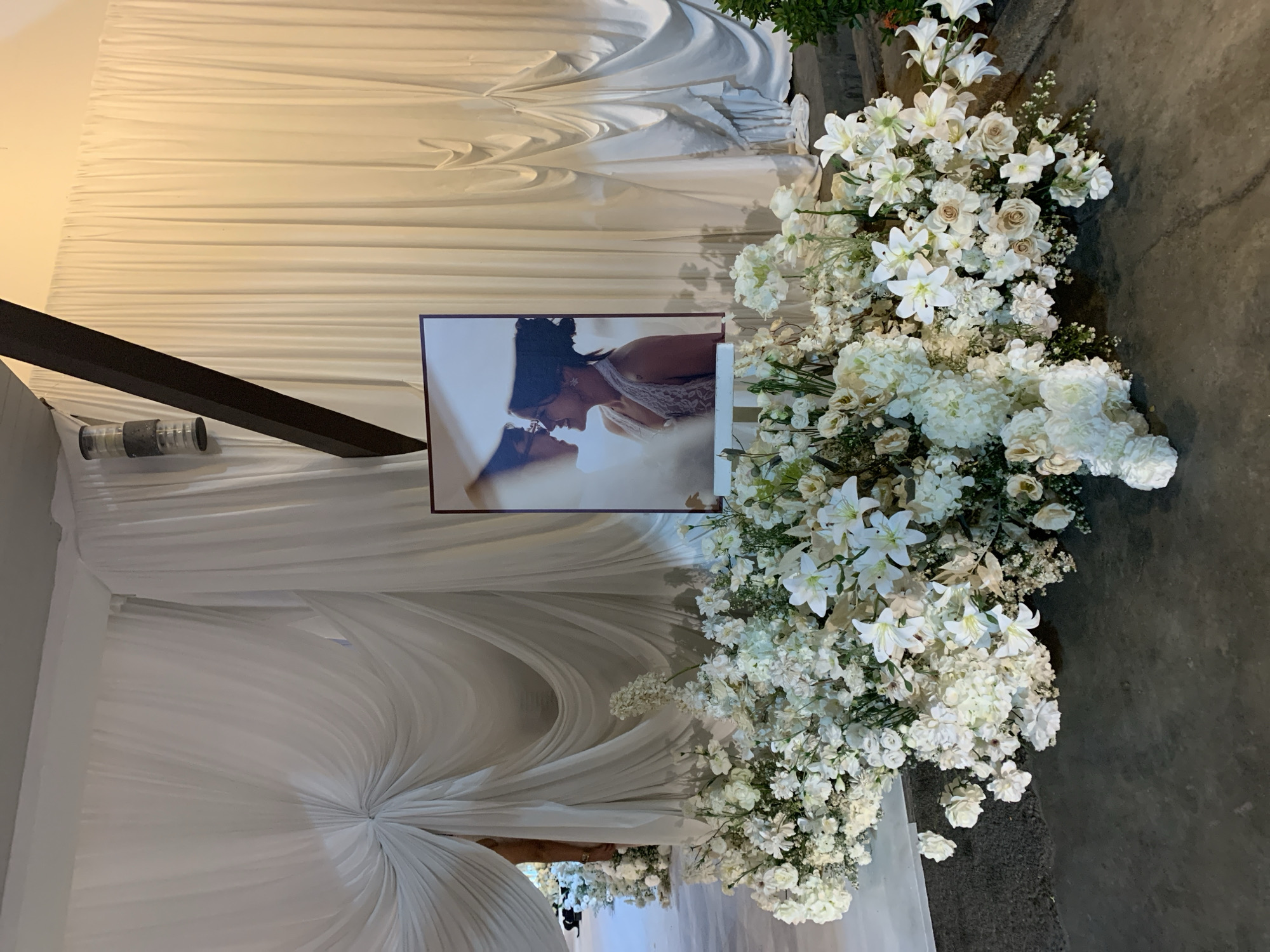 This image depicts a beautifully arranged setting, likely for a wedding or a significant celebratory event, taking place at บ้านบางรักษ์, Thailand. The scene is characterized by a romantic and elegant ambiance, primarily achieved through white decor and floral arrangements.
A prominent feature is a framed portrait of two individuals, presumably a couple, captured in an intimate pose with their faces close, appearing to kiss or share a tender moment. This photograph is centrally placed within the decorative setup.
Flanking the photograph and extending into the foreground are lush, cascading floral arrangements. These predominantly white flowers include white lilies, roses, and various other smaller white and cream blossoms, accented with hints of green foliage. They appear to rest on or spill over onto a textured concrete floor, which forms the ground surface in the scene.
The background is adorned with extensive white fabric draping, creating a soft, voluminous, and elegant backdrop. The fabric is artfully pleated and gathered, adding depth and texture to the setting. A dark, possibly wooden, structural beam runs diagonally across the upper left quadrant of the image, and a cylindrical, transparent object, possibly a light fixture or part of the decor, is visible beneath it.
The overall impression is one of sophistication and celebration. The lighting appears soft, contributing to the serene and romantic mood. There is no visible text in the image.