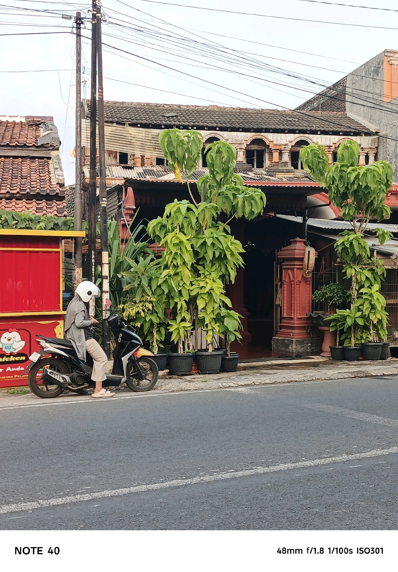 A person wearing a white helmet, grey cardigan, and light-colored pants is sitting on a black and yellow motorcycle parked on the side of a street. The motorcycle is positioned in front of a building with a red facade and a red sign displaying a cartoon chicken and the text "chicken" and "Terima Pelanggan Anda". Behind the motorcycle and building, there are lush green trees in black planters. The background features a two-story building with a tiled roof and decorative arched windows. Overhead power lines crisscross the sky. The scene appears to be in an urban setting in Kelurahan Cilacap, Kecamatan Cilacap, Indonesia.
