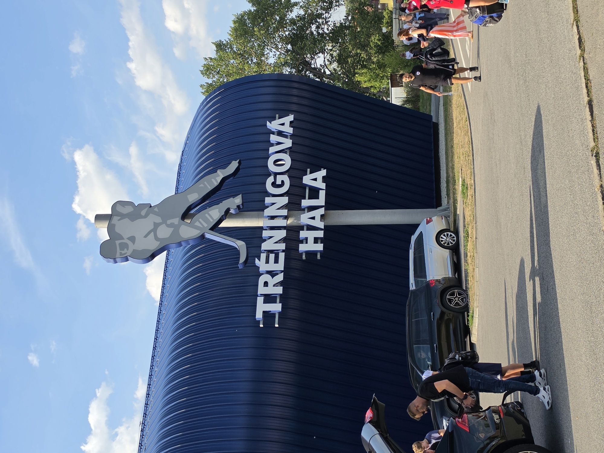 A wide shot captures the exterior of a blue, corrugated metal building with the text "TRÉNINKOVÁ HALA" in white, 3D lettering across its facade. A large, silver silhouette of a hockey player is mounted above the text. In the foreground, several people are seen near parked cars. On the left, two men are interacting with the open trunk of a black SUV, one appears to be putting something away. To the right of the building, a group of people are walking across a crosswalk. The sky is blue with scattered clouds, and trees are visible behind the building. The scene appears to be outdoors in daylight.