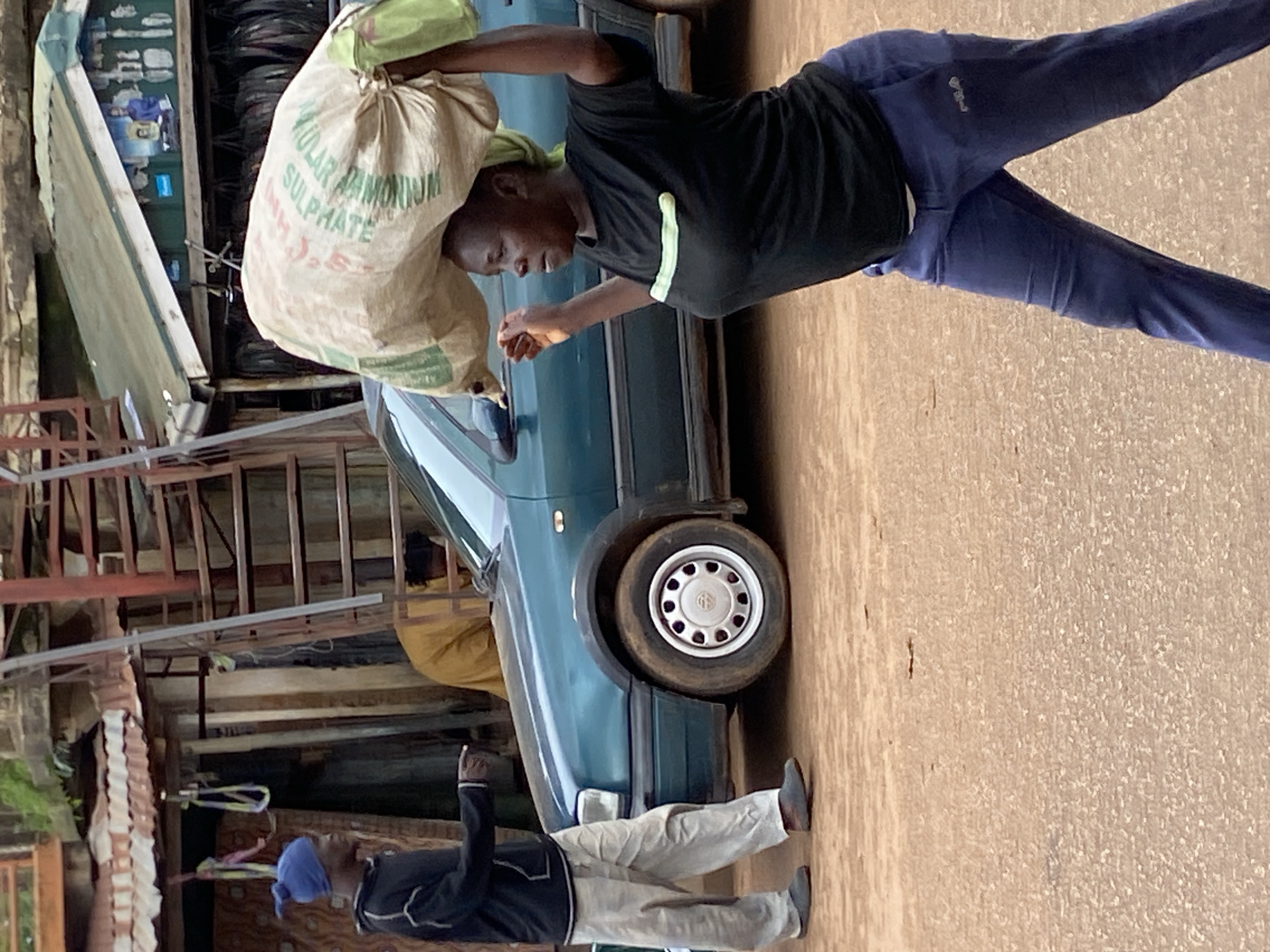 In Bukuru, Nigeria, a woman carries a large sack of goods on her head, walking past a teal sedan. She is wearing a black t-shirt with a light green stripe and blue athletic pants. A man in a blue head covering and black sweatshirt stands to the left, pointing towards the car. Behind them, a market stall with a corrugated metal roof displays various items, and a rusty metal staircase ascends to an upper level. The ground is a dusty, reddish-brown earth.