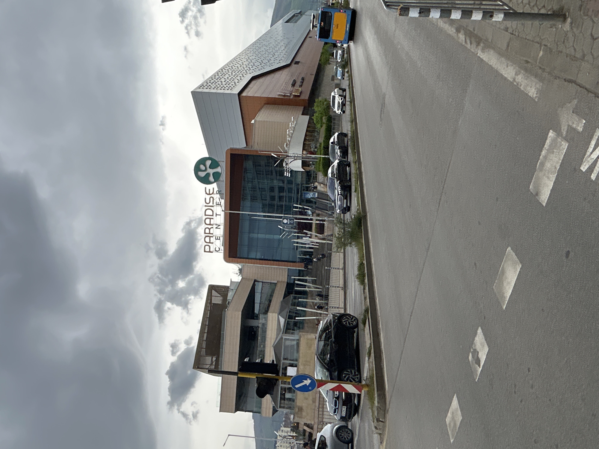 The image shows the Paradise Center shopping mall in Sofia, Bulgaria. The mall is located next to a road, with cars parked along the curb. A bus is visible further up the road. The sky is overcast. The shopping mall signage is in English. There are road markings on the road indicating traffic direction. A traffic light is also visible.