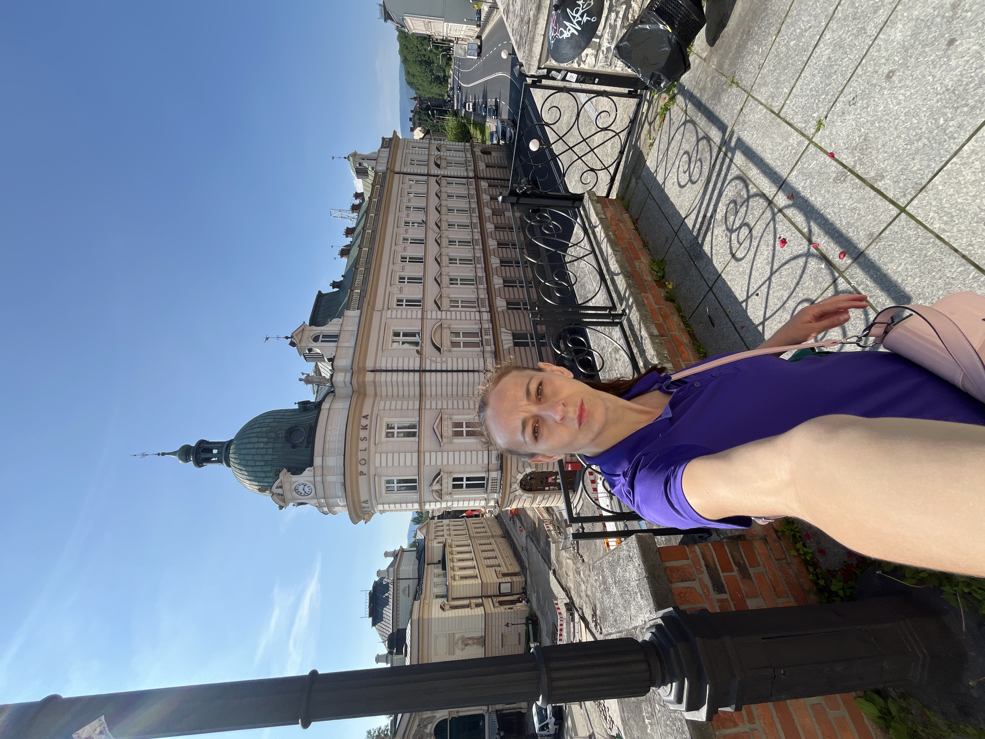 In a city scene with clear blue skies, a woman in a purple polo shirt takes a selfie from a stone-paved elevated walkway. She is positioned on the right side of the frame, holding her phone to capture the image. To her left, a black lamppost stands prominently. Behind her, a large, ornate building with a green dome and a clock tower dominates the view. The building features multiple windows and classical architectural details. In the background, a street lined with buildings and trees is visible, with cars driving on the road. The walkway is bordered by a low brick wall and a decorative black metal fence. The ground is paved with large square tiles, and scattered red petals are visible on the pavement, casting intricate shadows from the fence.