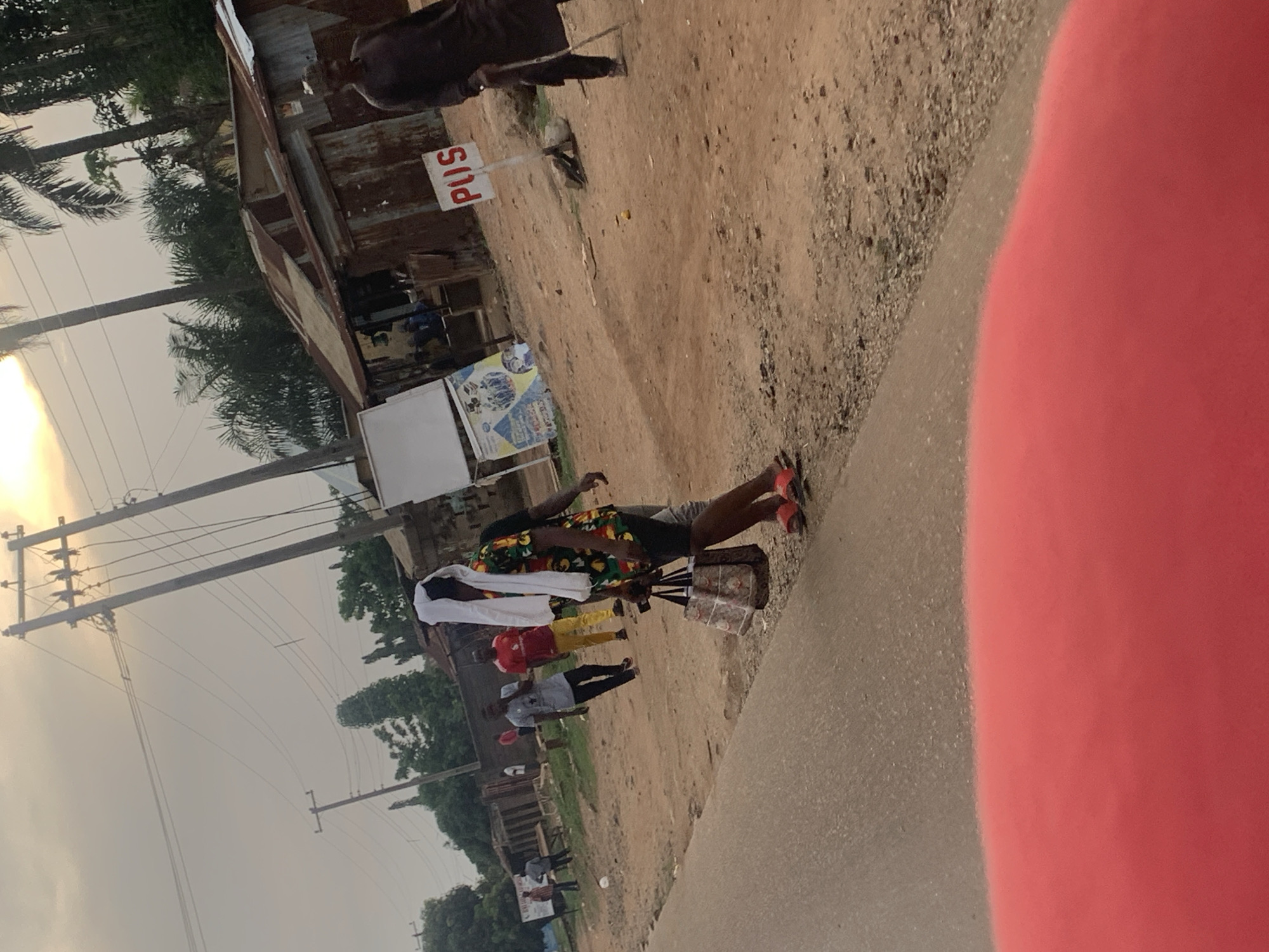This image, set in Agban, Nigeria, captures an outdoor street scene, likely during late afternoon or early morning, characterized by soft, diffused light and a low sun visible in the upper left. The setting features a paved road on the right and a wider, dusty unpaved path on the left, lined with rudimentary structures made of corrugated iron and wood, possibly indicating small shops or informal stalls.
The main subject is a person, likely a street vendor, standing by a small display cart on the edge of the paved road. They are wearing a colorful, patterned short-sleeved top (featuring green, red, and yellow hues), dark shorts, and distinct red open-toed sandals. A white cloth is draped over their head and shoulders. The cart beside them appears to hold various goods, though they are not clearly discernible.
Several other people are visible further down the dirt path, suggesting ongoing street activity. These include individuals in a red shirt and yellow pants, and another in a light blue/grey shirt. In the background, palm trees and other foliage are visible, and numerous utility poles with crisscrossing power lines span the sky.
A notable detail is a white sign with red lettering that reads "PUSS," mounted on a pole near one of the corrugated iron structures. The immediate foreground on the right side is partially obscured by a large, blurry red object. The scene portrays a typical, active roadside environment.