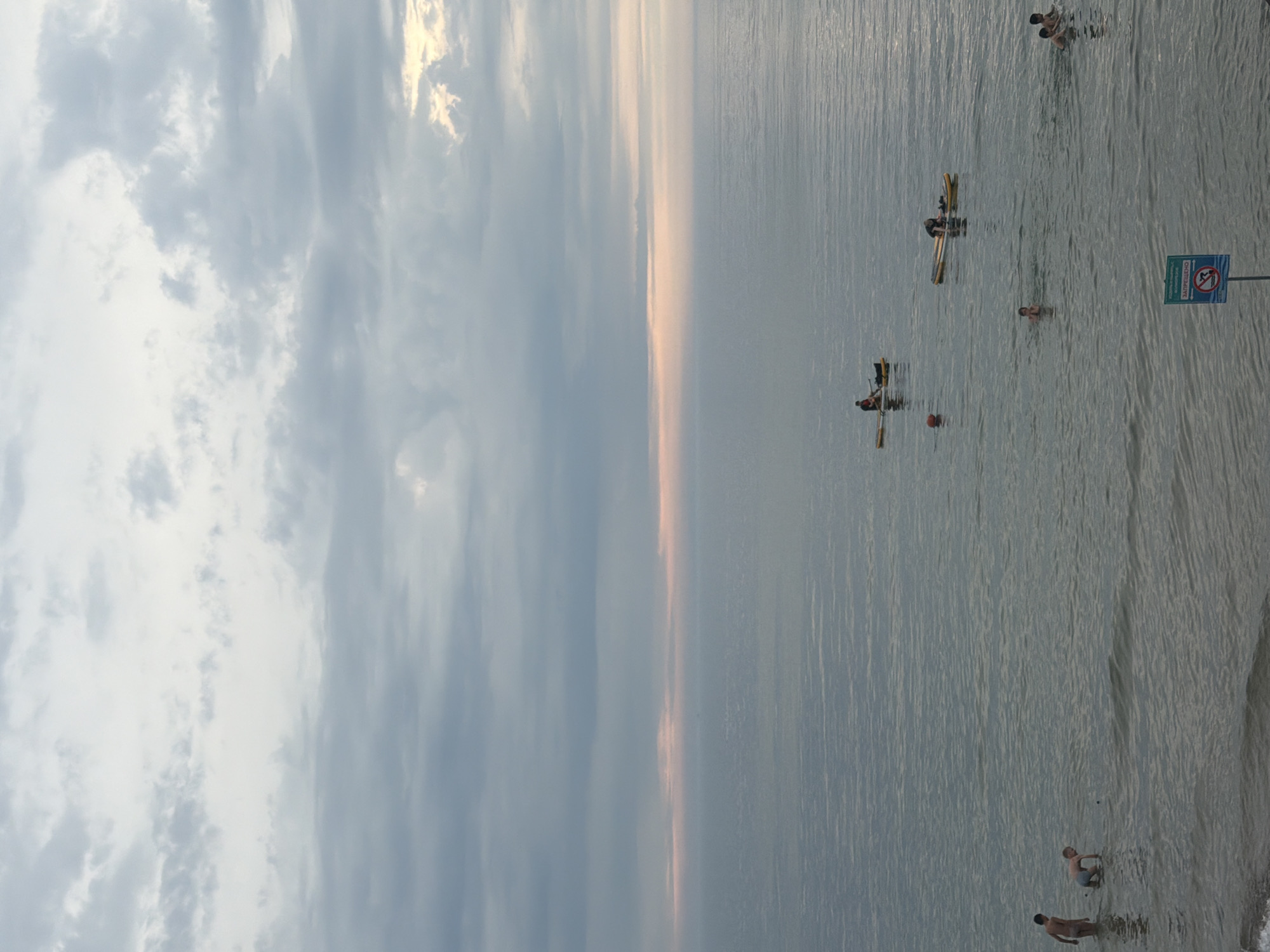 The image depicts a body of water, likely the Baltic Sea, with several people engaged in various water activities. In the foreground, two individuals are partially submerged in the water. Further out, a person is on a paddleboard, and another is in a kayak. The sky is overcast with clouds, and a hint of pink can be seen along the horizon, suggesting it might be dawn or dusk. The overall scene conveys a sense of a calm and recreational atmosphere. Given the context of Zelenogradsk, Russia, this scene is likely situated on the Zelenogradsk beach or a nearby coastal area.