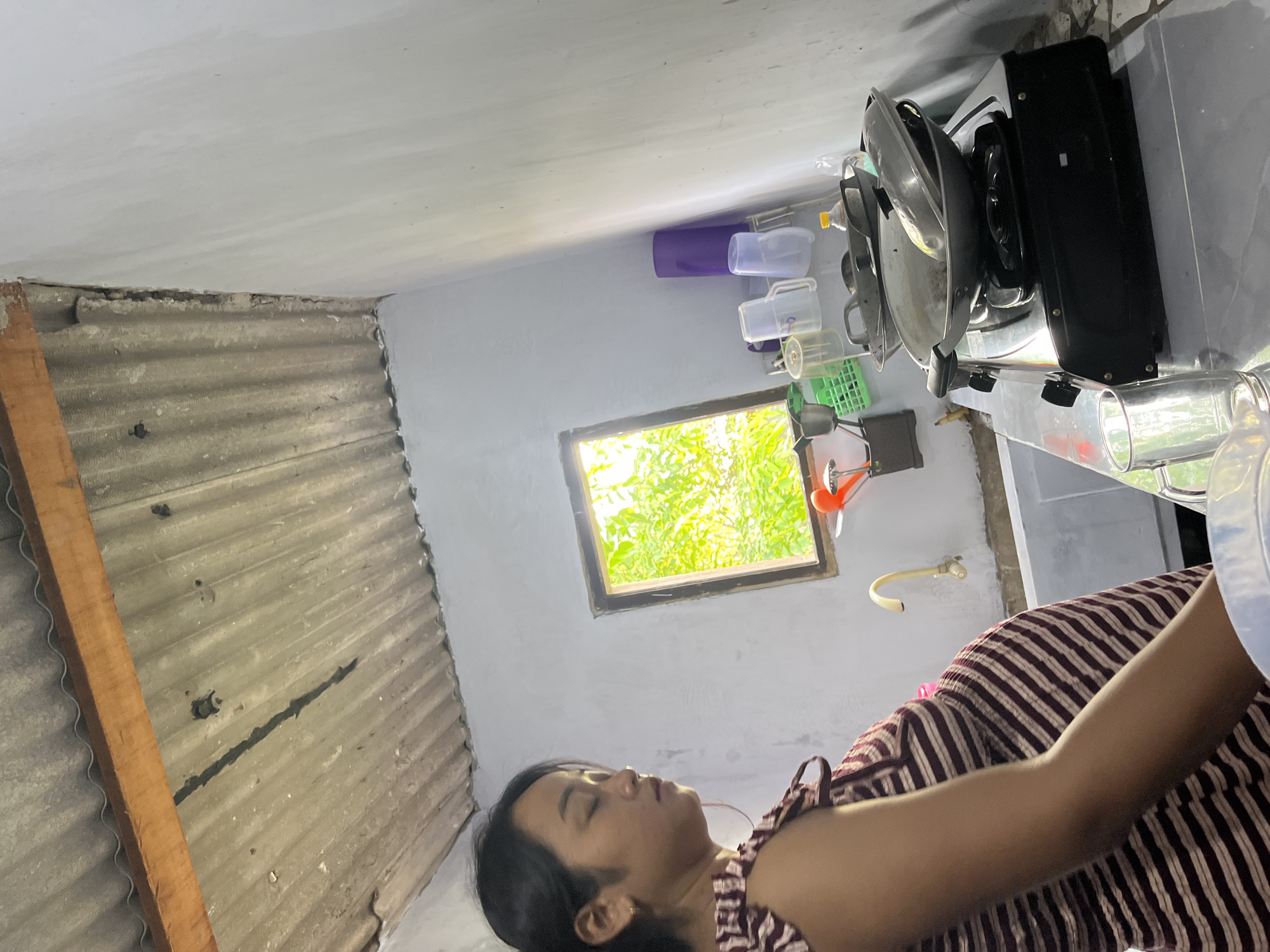 An Indonesian woman, appearing to be pregnant and wearing a striped dress, stands in a modest kitchen in Laban, Indonesia. She has her eyes closed and her head tilted downwards. The kitchen features a corrugated metal roof with visible wooden beams, white walls, and a small window looking out onto lush green foliage. A sink with a faucet is visible, as is a black gas stove with cookware on top. Several plastic cups and a purple cylinder are on a shelf. The overall environment suggests a simple, lived-in domestic space.