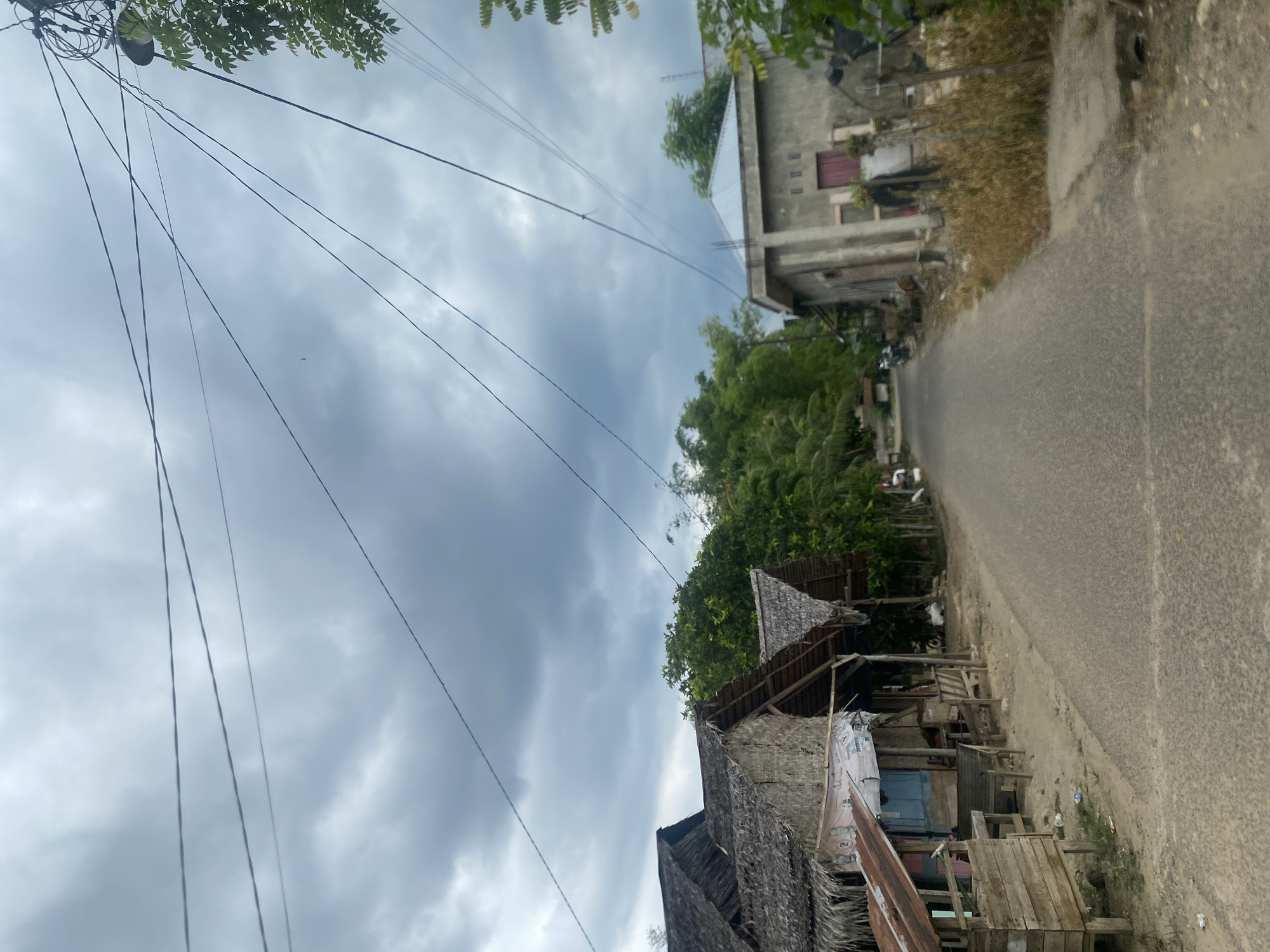 The image depicts a street in Kebonbaru, Indonesia. The street is paved and appears narrow. On one side of the street are buildings of varying construction styles. The closest building has a thatched roof and wooden supports. Further down the street is a more modern concrete building with a red door. Lush green vegetation is visible. Overhead, electrical wires stretch across the sky, which is filled with dark, cloudy skies.