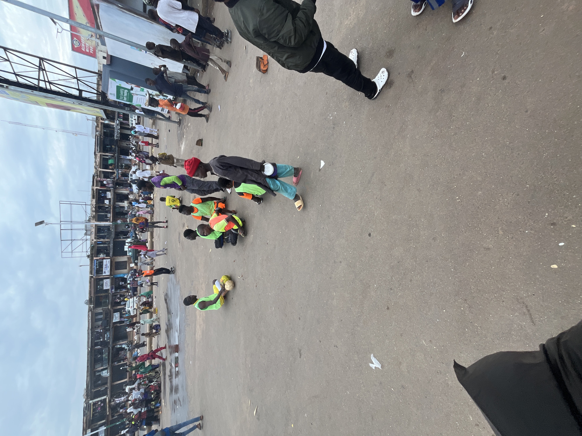 The image depicts an outdoor scene in Jos, Nigeria, possibly a market or a street with shops lining one side. The weather appears overcast. A group of people are present, with some in the foreground and others further back.
In the foreground, several individuals are kneeling or sitting on the ground. One person in a red cap and a dark jacket is holding a white bucket. Next to them, a person in a green and orange vest is kneeling, and another person in a similar vest is also on the ground, holding a soccer ball. Further to the right, another person in a green vest is kneeling.
In the background, there are more people, some standing and some seated. A prominent building with multiple stories and balconies runs along the side of the scene. Several signs are visible on the building. One large billboard on the left features red and white text, with "ALWAYS WITH YOU" partially visible. Another sign on a building displays green and white graphics with text that seems to be an advertisement. The ground is paved with asphalt and shows some signs of wear and tear, with scattered debris like paper. The overall impression is of a busy public space, possibly a gathering or a street performance involving some of the individuals. The time of day appears to be daytime, given the ambient light.