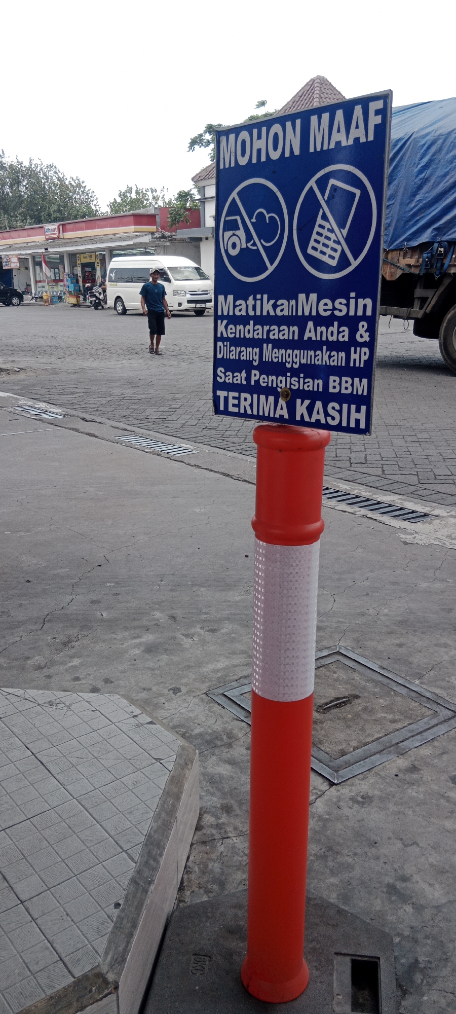 The image shows a street scene in Tebaloan, Indonesia. In the foreground, a red and white striped bollard stands next to a blue sign with white lettering. The sign reads "MOHON MAAF" (Please excuse) and displays two icons: one with a crossed-out vehicle and smoke, and another with a crossed-out mobile phone. Below these icons, the text continues: "Matikan Mesin Kendaraan Anda & Dilarang Menggunakan HP Saat Pengisian BBM TERIMA KASIH" (Turn off your vehicle engine & do not use HP while refueling thank you). In the background, a white van is parked near a minimarket with an "Indomaret" sign. A man in a dark blue t-shirt and black shorts is walking away from the van. A large truck with a blue tarp covering its load is visible on the right side of the image. The ground is paved with concrete and cobblestones, with visible cracks in the concrete. The sky is overcast.