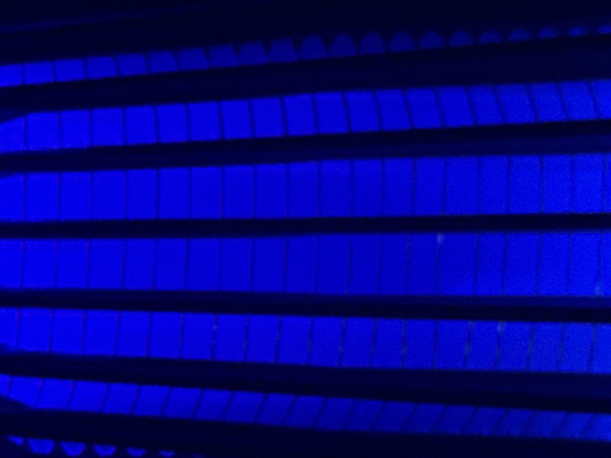 The image shows a close-up, abstract view of blue light filtering through vertical, dark, parallel structures. The lighting is intense and uniformly blue, creating a vibrant glow on the surfaces it illuminates. The structures themselves appear to be evenly spaced and form a pattern of repeating vertical elements with some horizontal segmentation within them. The overall impression is one of a rhythmic, perhaps artificial, lighting installation. There are no visible people or recognizable landmarks within the frame. The image does not contain any information to suggest a specific location in Makhachkala, Russia.