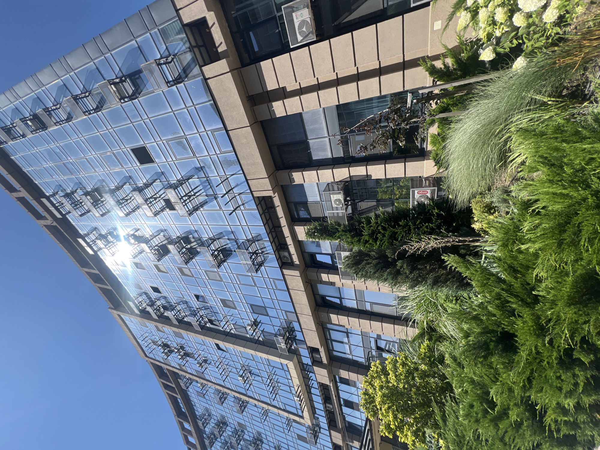 This low-angle, full-shot, outdoor, eye-level photograph captures a modern, multi-story building with a facade of blue glass windows and beige stone accents. The building is illuminated by bright sunlight, which reflects intensely off the glass. Numerous windows are open, with metal structures projecting outwards. Several outdoor air conditioning units are mounted on the exterior walls. In the foreground, lush green bushes and trees form a dense line, with a tall, slender evergreen tree on the right side of the frame. The sky above is a clear, bright blue. The image appears to depict a residential or commercial building in an urban setting, possibly Fontanka, Ukraine, characterized by contemporary architecture and well-maintained landscaping.