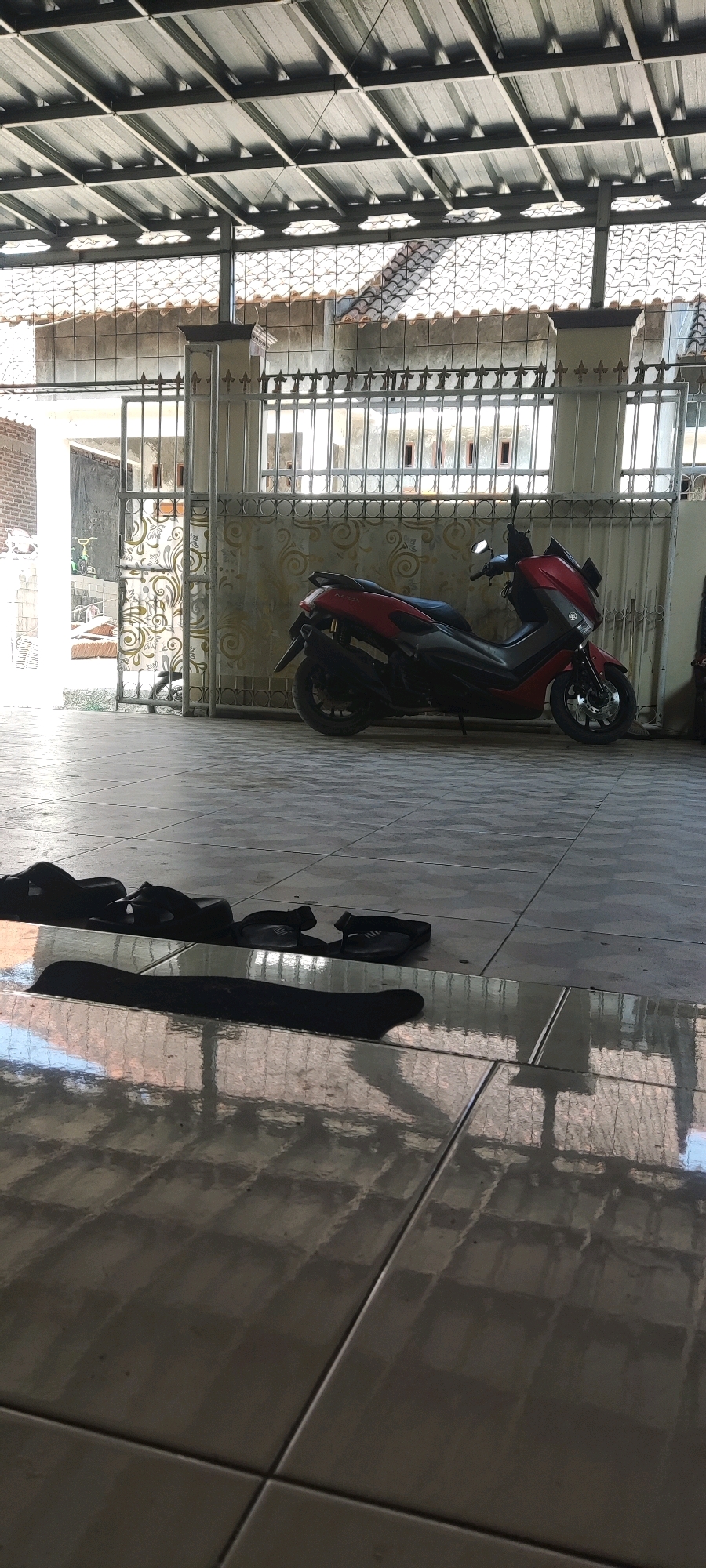 The image depicts an outdoor scene in Japura Kidul, Indonesia, likely a residential area. In the foreground, on a tiled floor that reflects the overhead structure, lie several pairs of black flip-flops. Further back, a red and black scooter is parked under a metal roof supported by beams, which is partially open to reveal a glimpse of a tiled roof and fence with ornate metalwork. Beyond the fence, residential buildings with tiled roofs are visible. The lighting suggests it is daytime, with sunlight casting shadows on the floor. The environment appears to be a covered patio or carport area.