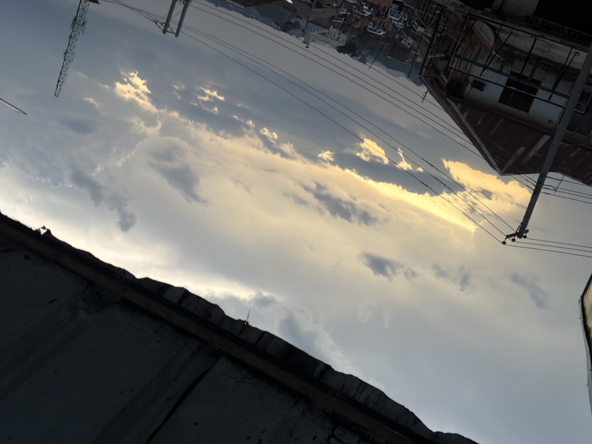 The image captures a street scene in Bukuru, Nigeria, under a dramatic sky. A low-angle shot looks up at a gray, cloudy sky, with streaks of yellow and orange light from what appears to be a sunset or sunrise breaking through the clouds. In the foreground, the edge of a dark, corrugated metal roof juts into the frame from the top. Below the sky, a two-story building with a rusty metal roof and balconies is visible on the left. Utility poles and numerous power lines crisscross the scene, obscuring the view of the sky and the street below. Further down the street, several vehicles, including cars and vans, are parked or in motion. A cellular tower stands tall on the right side of the image. The overall impression is of a busy urban environment transitioning into evening, with a visually striking sky.