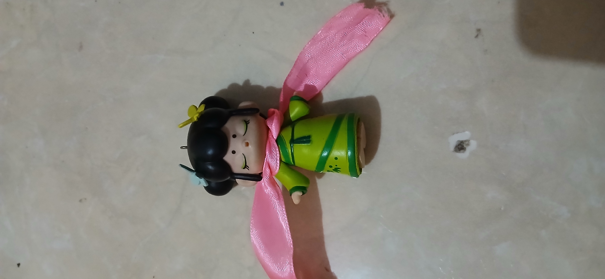 A small, green-robed doll with a pink scarf wrapped around its neck is positioned in the center of the frame. The doll has black hair styled in two buns, adorned with small light blue and yellow flowers. Its eyes are closed, and its expression is serene. The doll stands on a light-colored, textured surface, possibly a floor or tabletop, with scattered dark spots. The lighting suggests an indoor environment. The image was captured in Samarinda, Indonesia.