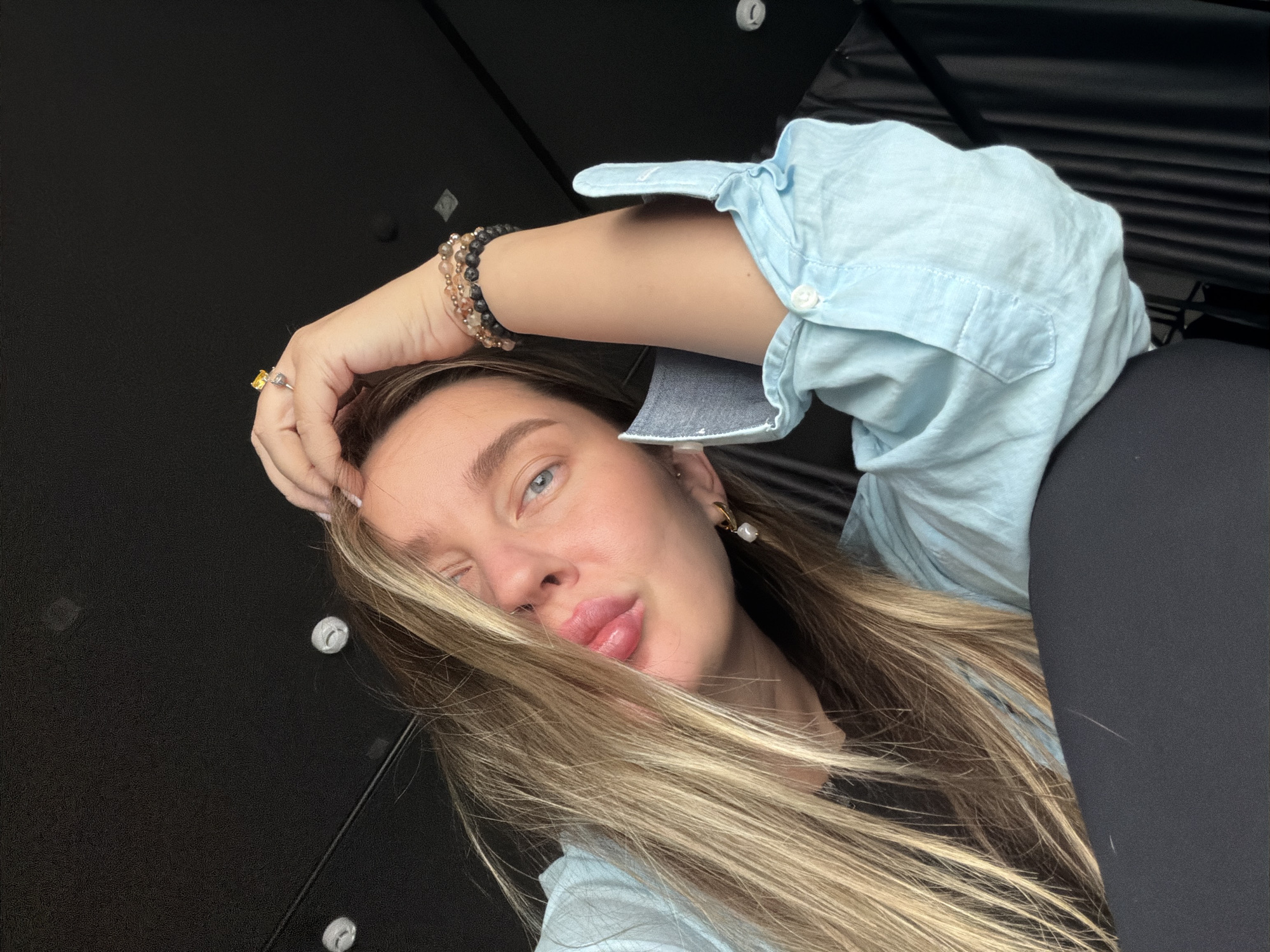 A young woman with long, blonde hair is reclining in what appears to be the interior of a car, possibly a taxi, with black leather seats visible on the right side of the frame. She is wearing a light blue button-down shirt and has multiple bracelets on her left wrist, which is resting near her forehead. She also has a ring on her left hand and a pearl earring in her left ear. Her face is slightly angled, with one eye open and the other closed in a wink. Her lips are slightly pursed. The background is a dark, ridged material, possibly the ceiling or roof of the car. The environment is indoor. It is not possible to determine the location of Moscow, Russia, from this image alone.