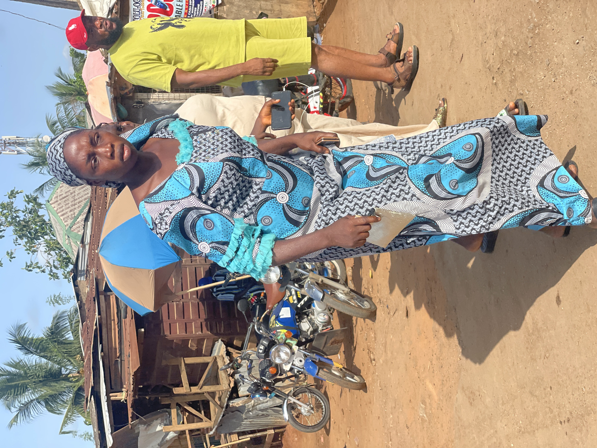 A woman in a blue and white patterned dress and headwrap is photographed at eye level in what appears to be a roadside setting in Ipota, Nigeria. She is holding a small, clear plastic bag in her left hand and has a somber expression. Behind her, partially obscured by a blue and brown umbrella, is a dilapidated wooden structure with a corrugated metal roof. Two motorcycles are parked in front of the structure. A man wearing a yellow shirt and shorts, and a red baseball cap is standing in the background. Another person is lying on a light colored mattress or blanket between the woman and the man. The ground is dirt, and there are palm trees in the background.