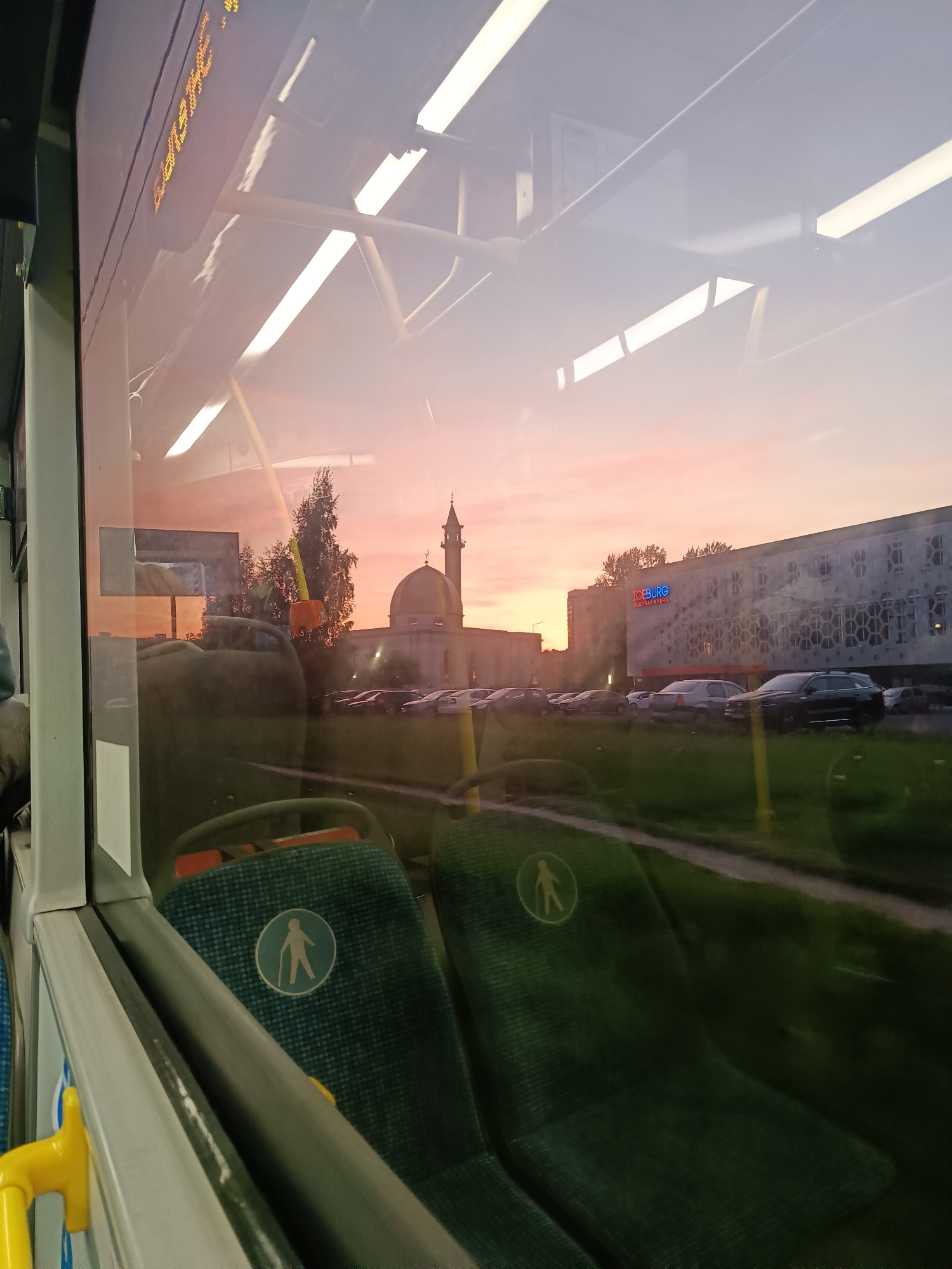 The image, taken from inside a bus, shows a view through the bus window. Two green seats with symbols indicating priority seating are visible in the foreground. Beyond the window, a street scene unfolds at sunset. A large, light-colored building with a dome and a minaret, likely a mosque, stands prominently in the middle ground. To the right of the mosque, a modern building with a sign "ICEBERG" in Cyrillic script is visible. Several cars are parked in a lot in front of the buildings. The sky is colored in shades of pink and orange, indicating sunset. The reflection in the bus window captures elements of the bus interior, including ceiling lights and a digital display. The image is likely taken in Saint Petersburg, Russia, given the Cyrillic script on the "ICEBERG" building, which is the "Iceberg Skating Palace" located there.