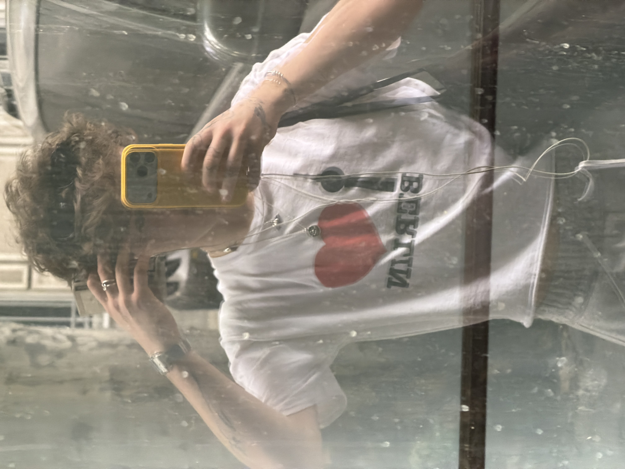 The image appears to be a reflection in a dirty window or other reflective surface. The reflection shows a person with curly hair holding a yellow phone, taking a selfie. The person is wearing a white shirt with a red heart and reversed black text that reads "I <3 BERLIN." The person is also wearing a watch and bracelets. Parts of a car and the street are visible in the background. There is no reliable evidence in the image that indicates this is Belgrade, Serbia. Therefore, a specific location in Belgrade cannot be determined.
