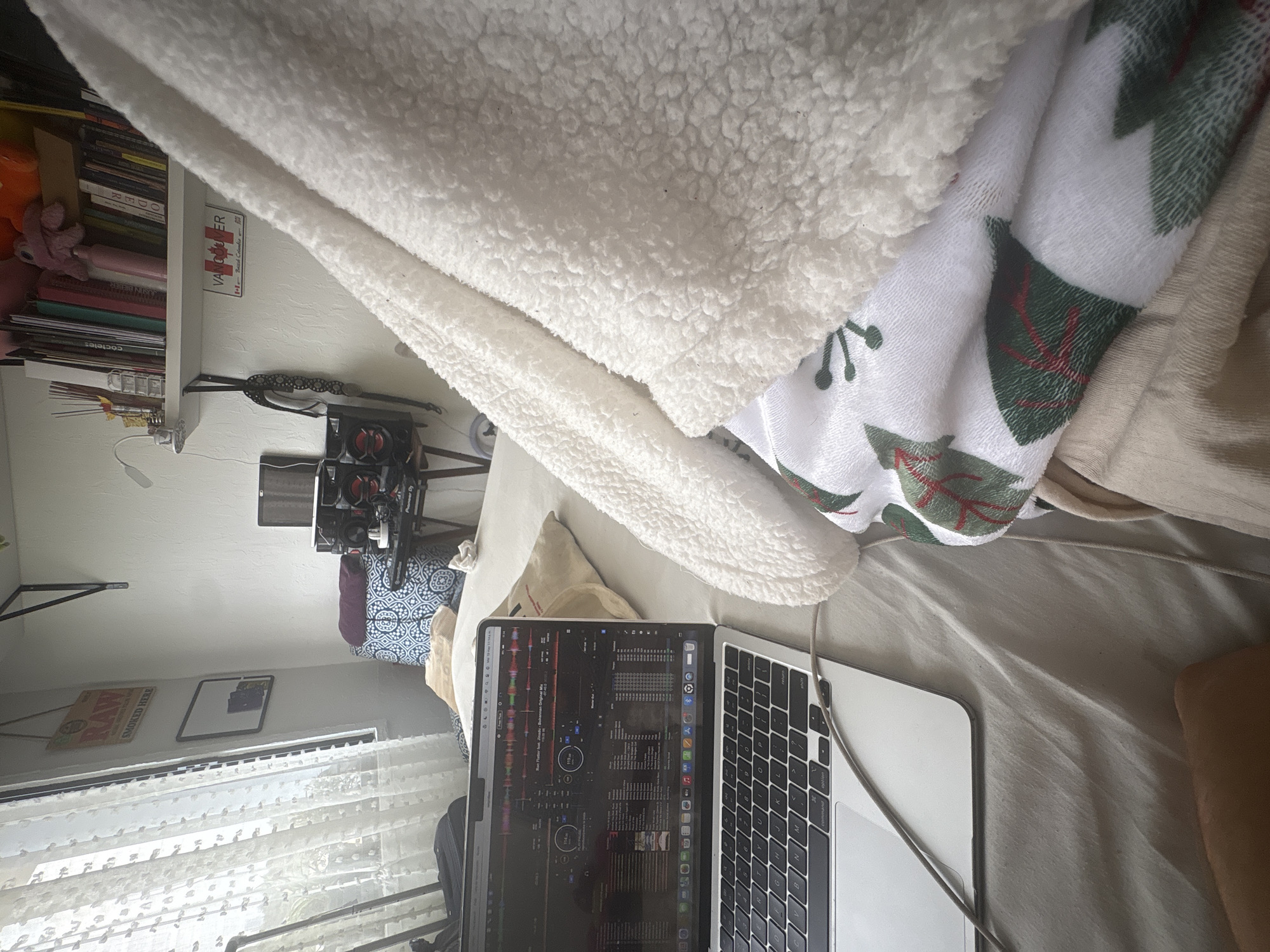 The image shows a laptop open on a bed, partly covered by a white fluffy blanket and another patterned blanket with green tree designs. A white bookshelf with books and a "Vancouver" sign are visible in the background. A camera setup stands nearby, suggesting a filming or photography workspace. Near the window, there is a "RAW Smoked Here" sign hanging above it, the curtains are sheer and white. The overall setting appears to be a room used for both work and relaxation.