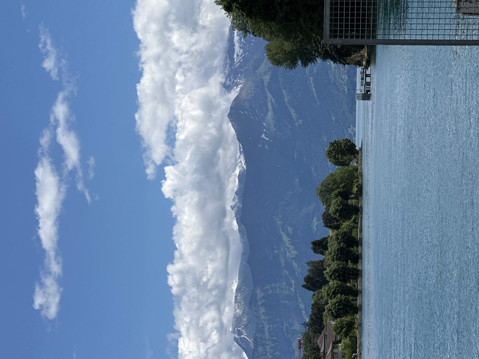 A wide shot captures a scenic view of Lake Thun, Switzerland, on a bright, partly cloudy day. The foreground is dominated by the tranquil, turquoise water of the lake. In the middle ground, a verdant shoreline stretches across the image, lined with mature trees. A row of buildings, appearing residential, is nestled behind the trees on the left side of the frame. Further out on the lake, a small pier extends from the shore, with a white structure on its end. A thin, dark line of vegetation borders the far side of the lake. The background is defined by a majestic mountain range, with the peaks dusted with snow. Lush green slopes lead up to the rugged, rocky summits. The sky above is a vibrant blue, dotted with fluffy white clouds. A metal fence encloses the right side of the foreground.