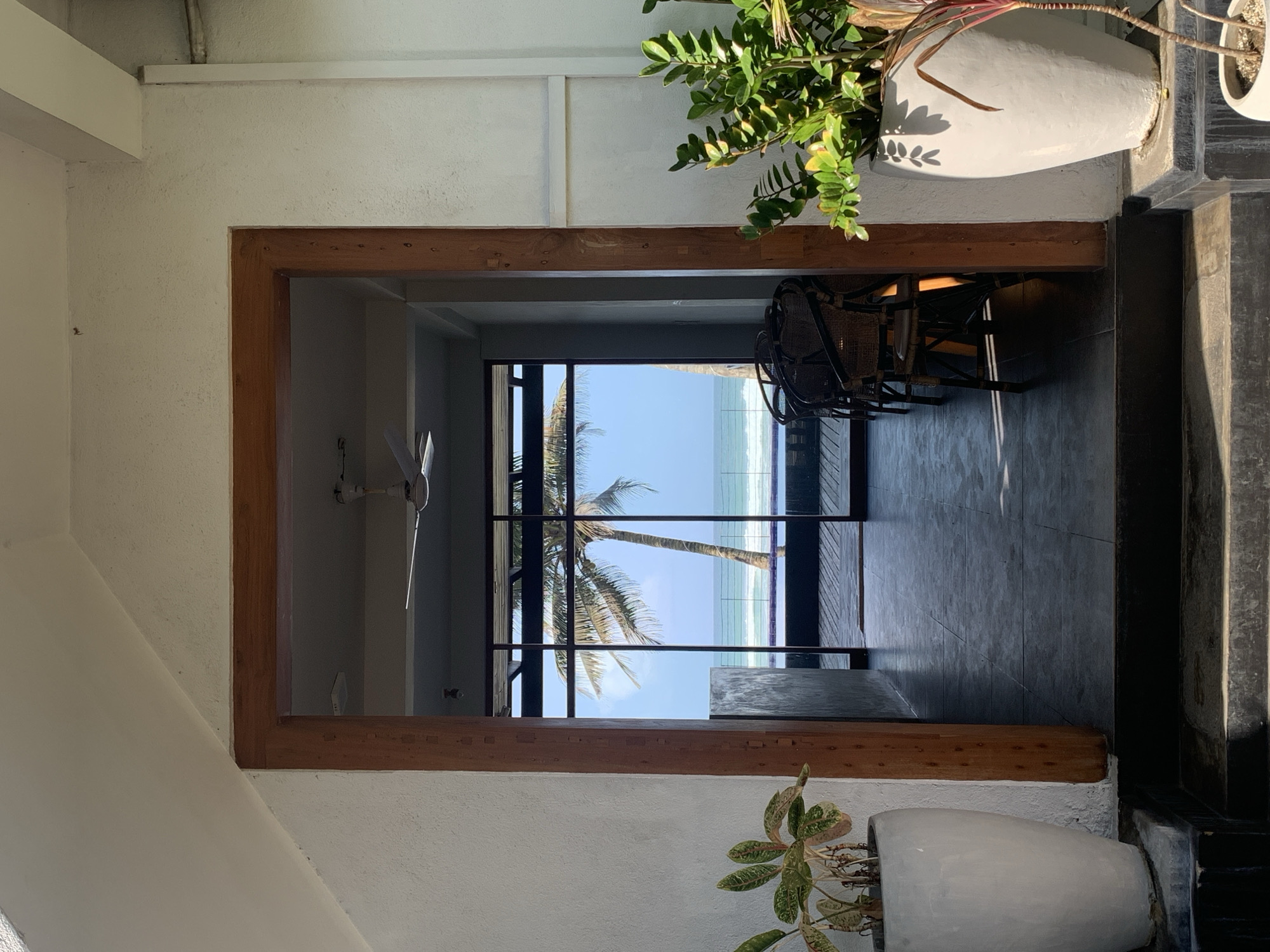The image shows an interior view of a space in Ahangama, Sri Lanka, framed by a wooden doorway. Through the doorway, a palm tree is visible, suggesting a location near the beach. The ocean and blue sky are visible between the branches of the palm. Inside the space, a ceiling fan is noticeable on the left. To the right of the doorway, a wicker chair and table are visible. The lighting suggests a daytime scene. Two potted plants sit on either side of the doorway. The floor appears to be dark grey stone or tile. The overall setting is a coastal building with an open view of the beach.
