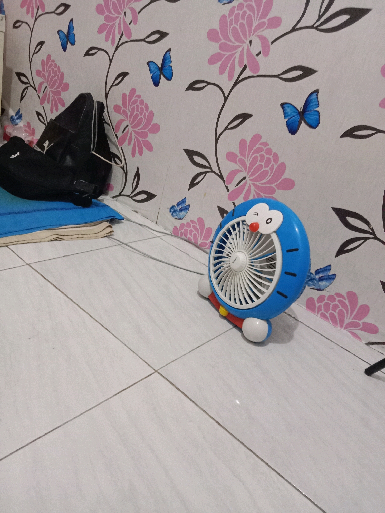 A blue Doraemon fan is on a white tiled floor. The fan has white blades and is shaped like Doraemon's head with a red nose and white eyes. It has white feet and a yellow accent on its body. The background is a white wall with a pink floral and blue butterfly pattern wallpaper. To the left of the fan, there are black bags and some blue and tan fabric items stacked on the floor. The scene appears to be indoors, in an unidentifiable location.