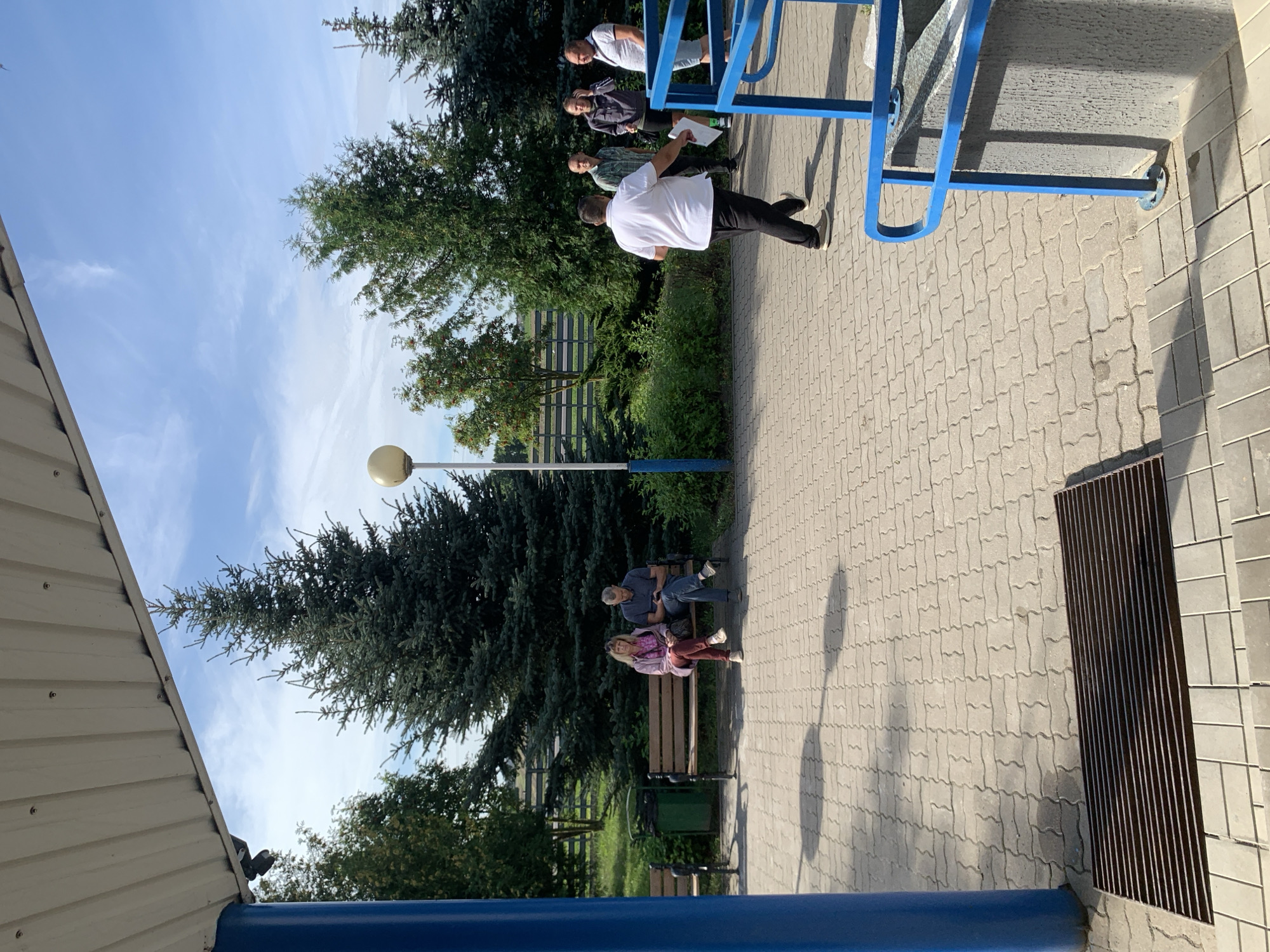 In Бокшыцкі сельскі Савет, Belarus, a group of people are visible outdoors on a paved area under a bright, cloudy sky. In the foreground, a blue metal railing frames the lower right of the scene, with a metal grate on the ground to the left. A man in a white shirt and black pants is walking away from the camera towards the right. Further back, two people, a woman and a man, are seated on a park bench. The woman has blonde hair and is wearing a pink jacket and burgundy pants, while the man is wearing a blue shirt and dark pants, looking at his phone. To their left is a green trash can. Behind the bench, several large evergreen trees and other foliage are present. In the background, a group of three men are standing together, and another evergreen tree is visible. The paved ground is made of interlocking bricks.