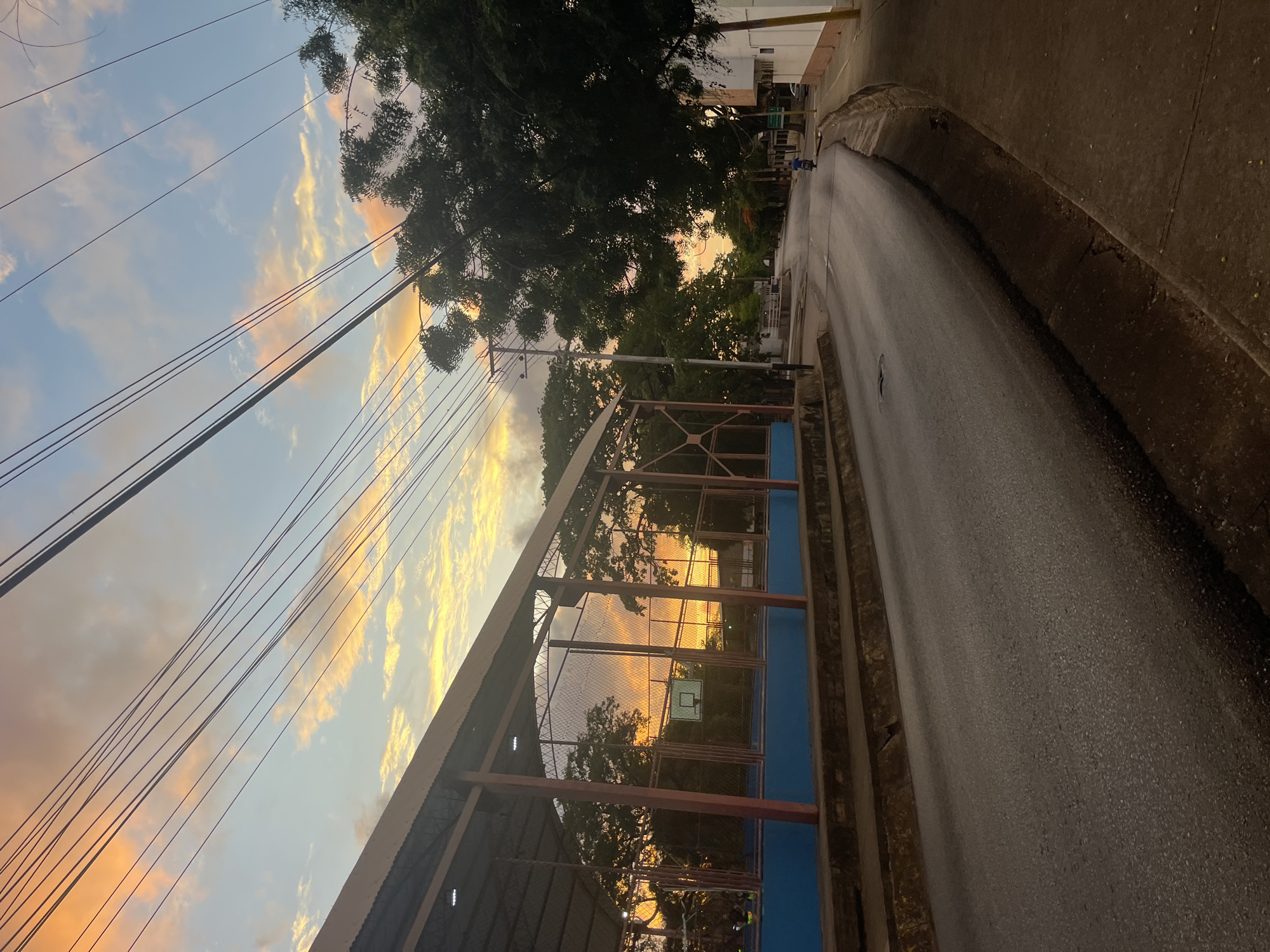 The image depicts a street scene in Santa Ana, Venezuela, during a sunset. A basketball court with a covered roof is visible on the left, featuring a basketball hoop and net. Overhead power lines crisscross the sky, which is painted with hues of orange, pink, and blue from the setting sun. Trees line the right side of the street, with a few buildings visible in the background. The asphalt road curves gently, with a concrete sidewalk and drainage ditch on the right. A lone motorcyclist is seen riding away from the viewer down the street. The overall impression is of a quiet, late afternoon in a residential area.