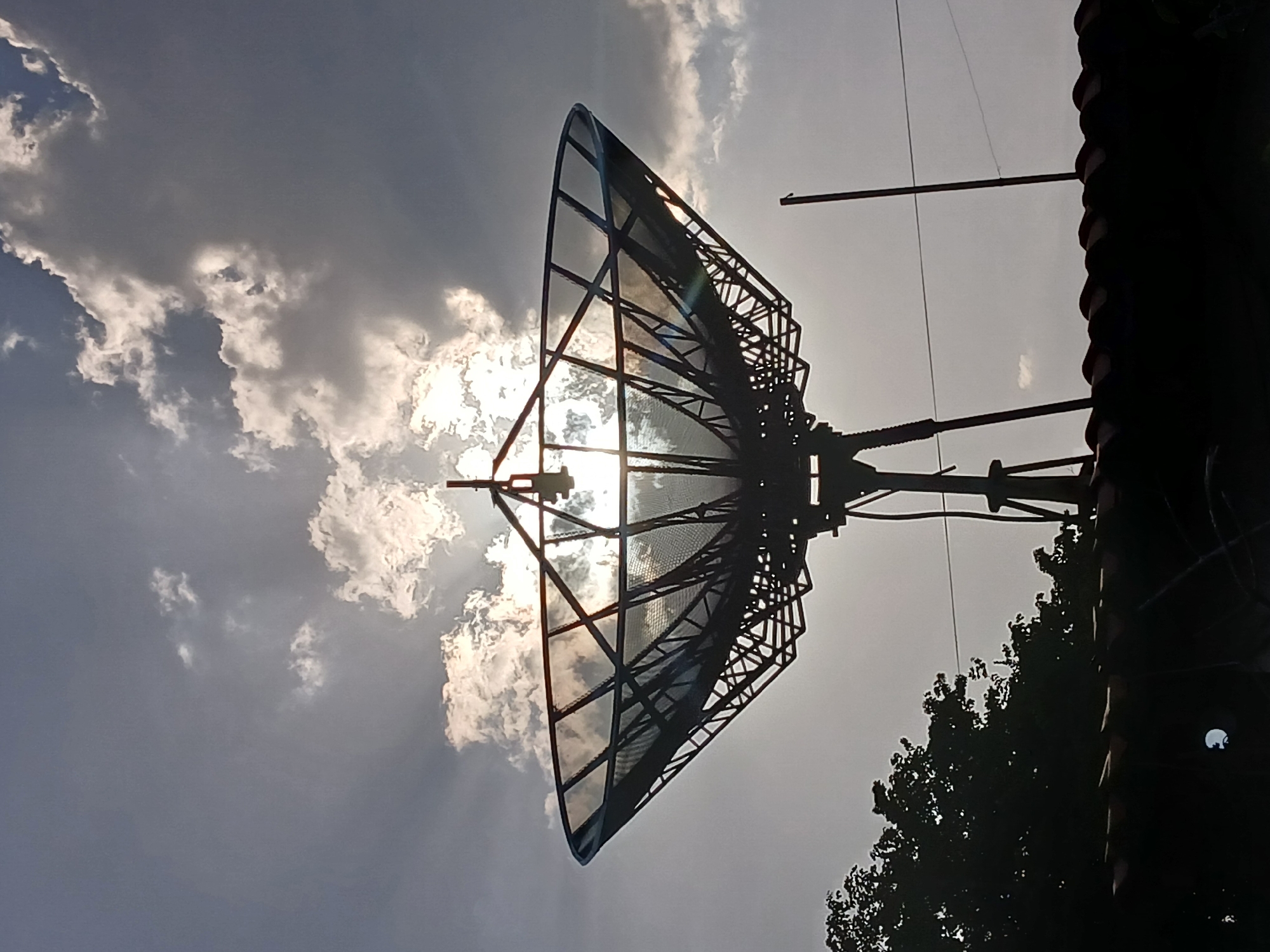 A low-angle shot depicts a large satellite dish against a sky with scattered clouds. The sun shines through the dish's mesh surface, creating a bright glare. The surrounding environment appears partially obscured by a dark structure, possibly a roof or building, and a tree. Specific details regarding the location in Ciudad Guayana, Venezuela are not apparent from the image.