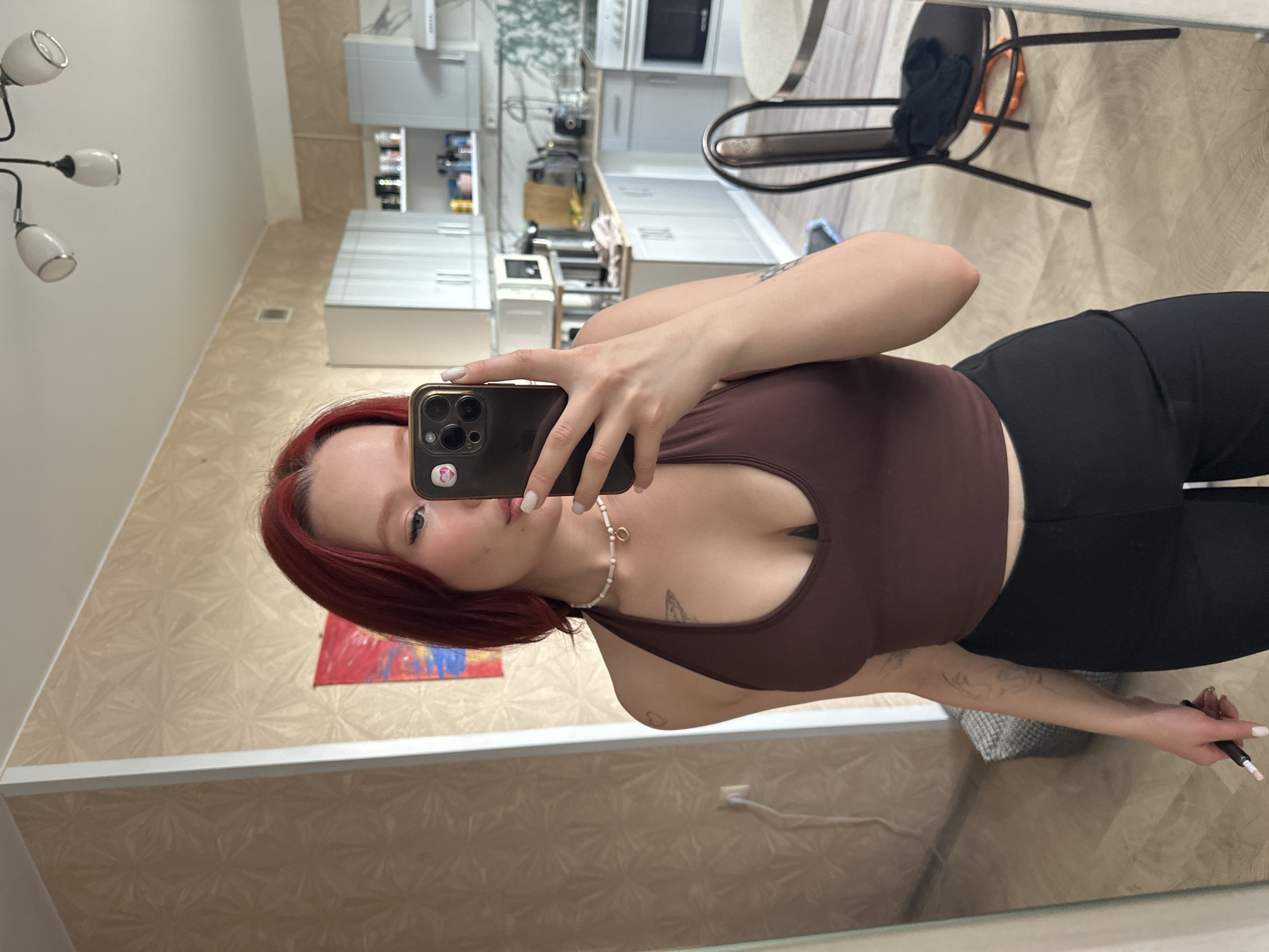 A young woman with red hair is taking a mirror selfie in what appears to be a kitchen area of a residential apartment. She is wearing a brown halter-neck top and black pants. She is holding a smartphone in her right hand, which is positioned to capture the reflection. A painting is visible on the wall behind her. The kitchen features white cabinets, a microwave, a stove, and various items on shelves. In the foreground, a chair is visible with some clothing on it. The floor is tiled. The overall setting suggests an indoor, domestic environment.