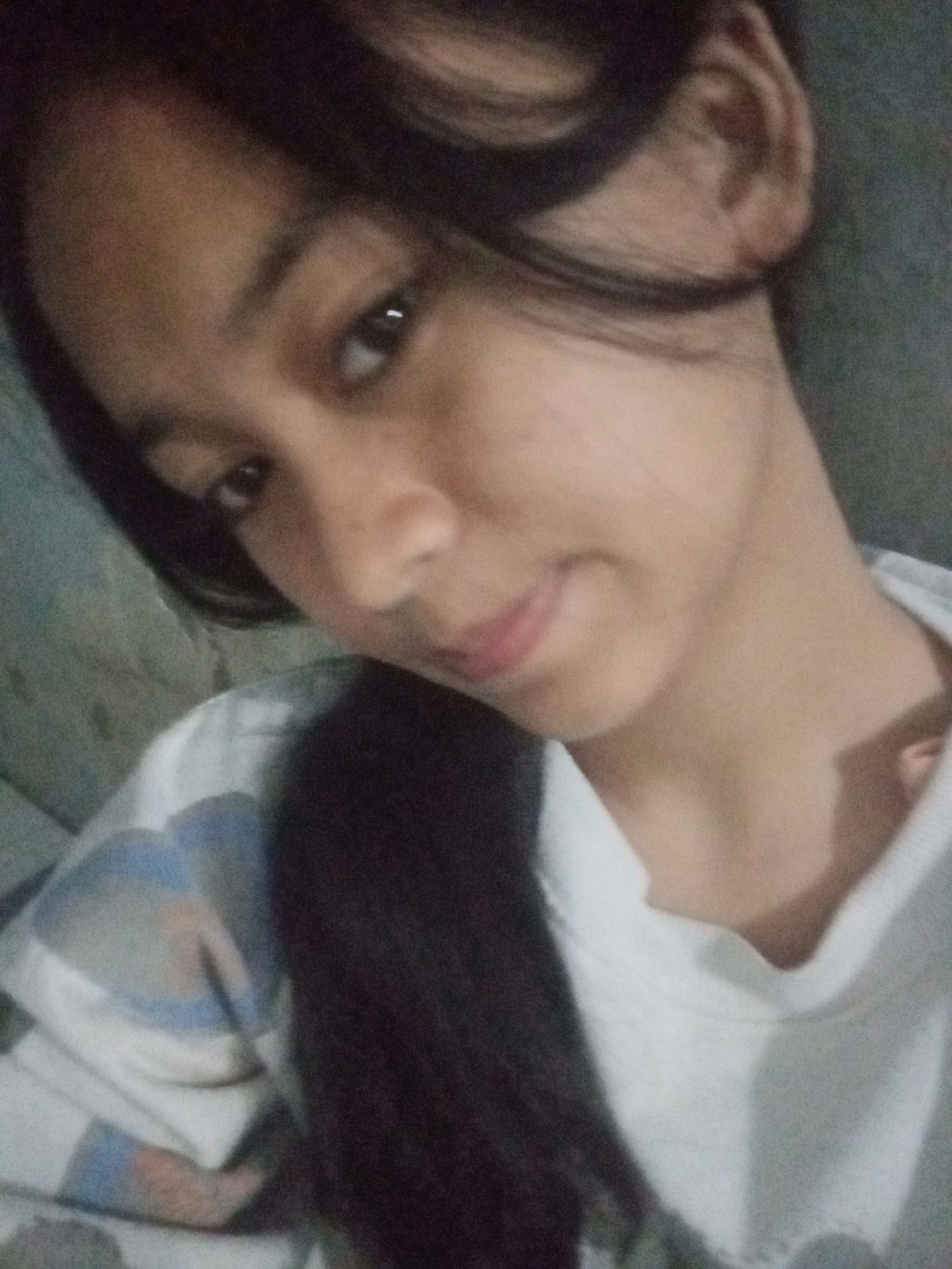 A young woman of East Asian descent is pictured in a close-up, selfie-style shot. She has dark hair that falls around her face and shoulders, and her skin tone is light to medium brown. Her eyes are dark and appear to be looking slightly to the left of the camera. Her lips are slightly parted, and she is wearing a white top with a blue and gray patterned design on the collar area. The background is dark and out of focus, suggesting an indoor setting. There is no discernible information within the image to determine the specific location in Badas, Indonesia.