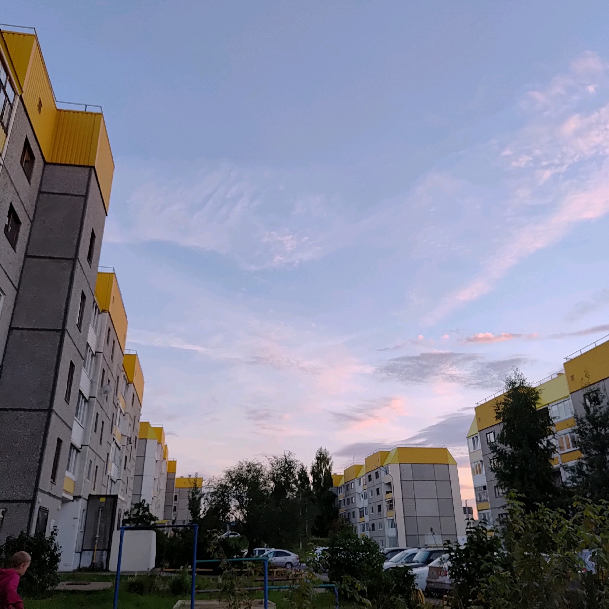 The image shows a residential area in the city of Markovsky, Russia, under a soft, twilight sky. Several multi-story apartment buildings, characteristic of Soviet-era architecture with grey concrete exteriors and bright yellow roofs, dominate the scene. The buildings are arranged in a way that suggests a courtyard or communal living space between them. Some windows in the buildings are visible, indicating occupied residences.
In the foreground, a small child wearing a pink hoodie is partially visible on the left side of the frame, facing away from the camera and towards the buildings. A low-lying green area with grass and some bushes surrounds the base of the buildings, suggesting a park or recreational space. A blue and white structure, possibly part of a playground or a fence, is present in the lower-left quadrant.
Several cars are parked in front of the buildings, contributing to the urban residential environment. The sky is a gradient of soft blues and pinks, with wispy clouds, suggesting either sunrise or sunset. The overall atmosphere is one of quiet residential living in a slightly aged urban setting, with the natural light of the transitioning day adding a serene quality to the scene.