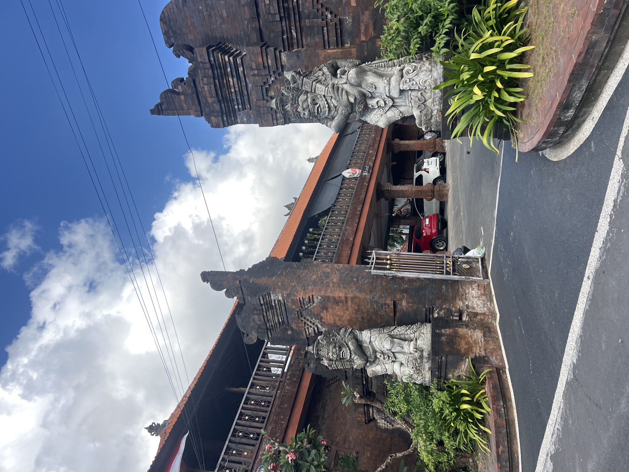 The image depicts the entrance to a building in Denpasar, Indonesia. Two large stone statues of gargoyle-like figures guard the entrance, which is made of weathered brick. The building itself features a multi-story structure with a wooden balcony and a terracotta tiled roof. Several cars are parked in the driveway, and lush green foliage surrounds the entrance. The sky above is a vibrant blue with scattered white clouds, and power lines are visible across the top of the frame.