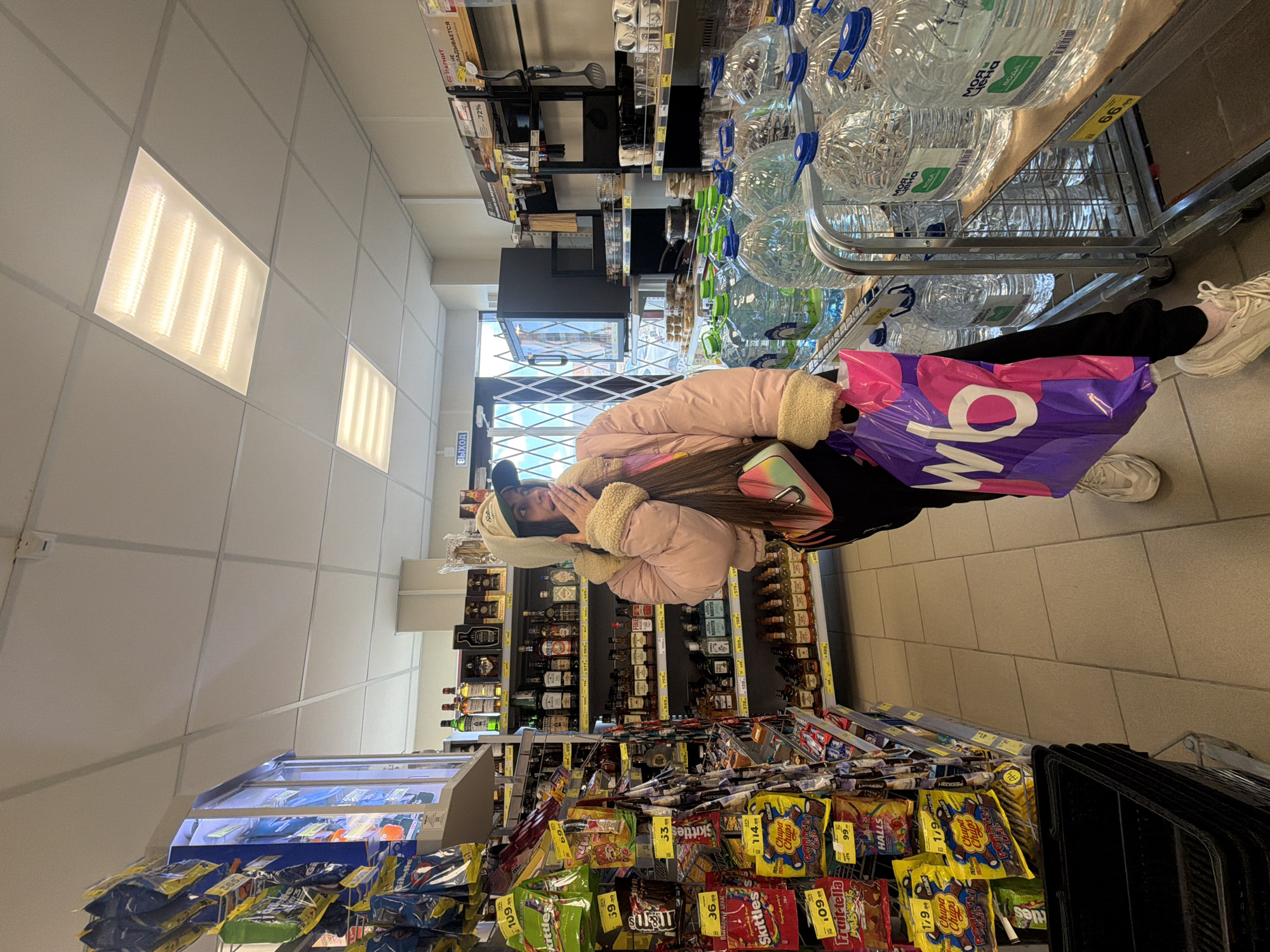The image shows a young woman in a supermarket aisle, likely in Saint Petersburg, Russia, based on the Cyrillic text on the products and signage. She is wearing a pink jacket with a fur-lined hood and carrying a purple shopping bag with white lettering. The aisle is stocked with various goods, including bottled water, liquor, and confectionery. The woman is positioned between shelves of bottled water and shelves of alcoholic beverages. The lower part of the frame showcases an assortment of candies and snacks, with visible brands such as Skittles and Chupa Chups. The lighting in the supermarket is bright, with visible panel lights on the ceiling. The overall scene depicts a mundane activity of grocery shopping.