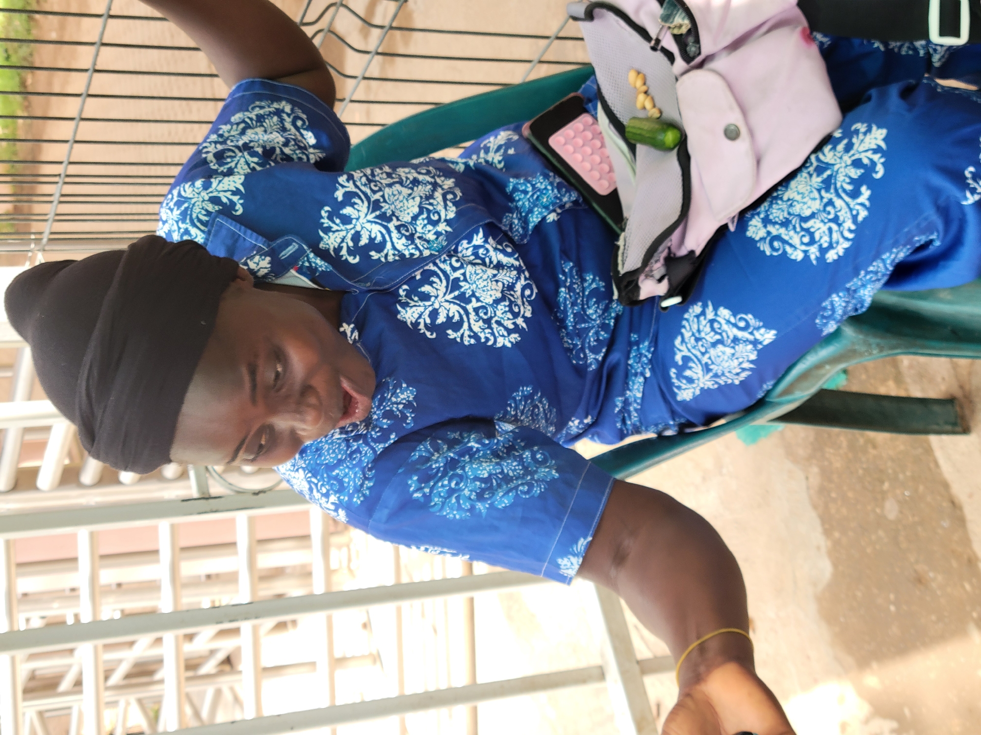 A medium shot depicts a middle-aged woman wearing a patterned blue shirt and a black head covering, sitting on a green chair outdoors. A light purple bag rests on her lap, which also contains a black phone and what appears to be a cucumber. The scene is set against a background of a white railing and what appears to be an orange brick surface. The specific location in the city of Abuja, Nigeria cannot be determined from the image.