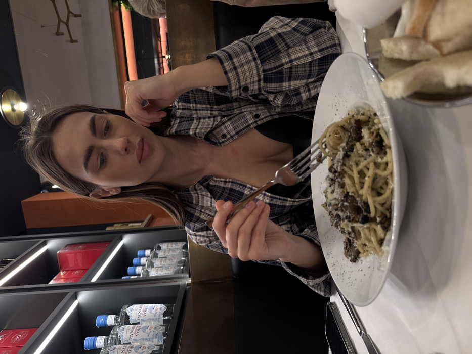 The image depicts an adult female dining in an indoor restaurant, specifically identified as PANAIR Ristorante, located at Via Sottoriva, 18, 37121 Verona VR, Italy. The female, appearing to be in her 20s or 30s, has long, straight, dark brown hair and is dressed in a plaid button-up shirt (black, white, and tan pattern) worn open over a black undergarment. A faint reddish mark is visible on her upper chest. She is seated at a white table, holding a silver-colored fork with her right hand, poised above a white plate containing spaghetti adorned with a dark, finely chopped ingredient (consistent with mushrooms or truffle), grated cheese, and black pepper. Her left hand rests near her chin, and her gaze is directed downward towards her meal. To her left on the table, a black smartphone with a silver edge and a knife are visible. Partially visible to her right is a metal basket containing pieces of light-colored bread. The background features a black shelving unit with integrated LED strip lighting, displaying multiple bottles of "PANAIR VODKA" with blue and white labels, and red boxes on upper shelves. The overall environment is dimly lit, suggesting an evening setting, with an orange-brown wall panel and a circular ceiling light fixture in the background.
