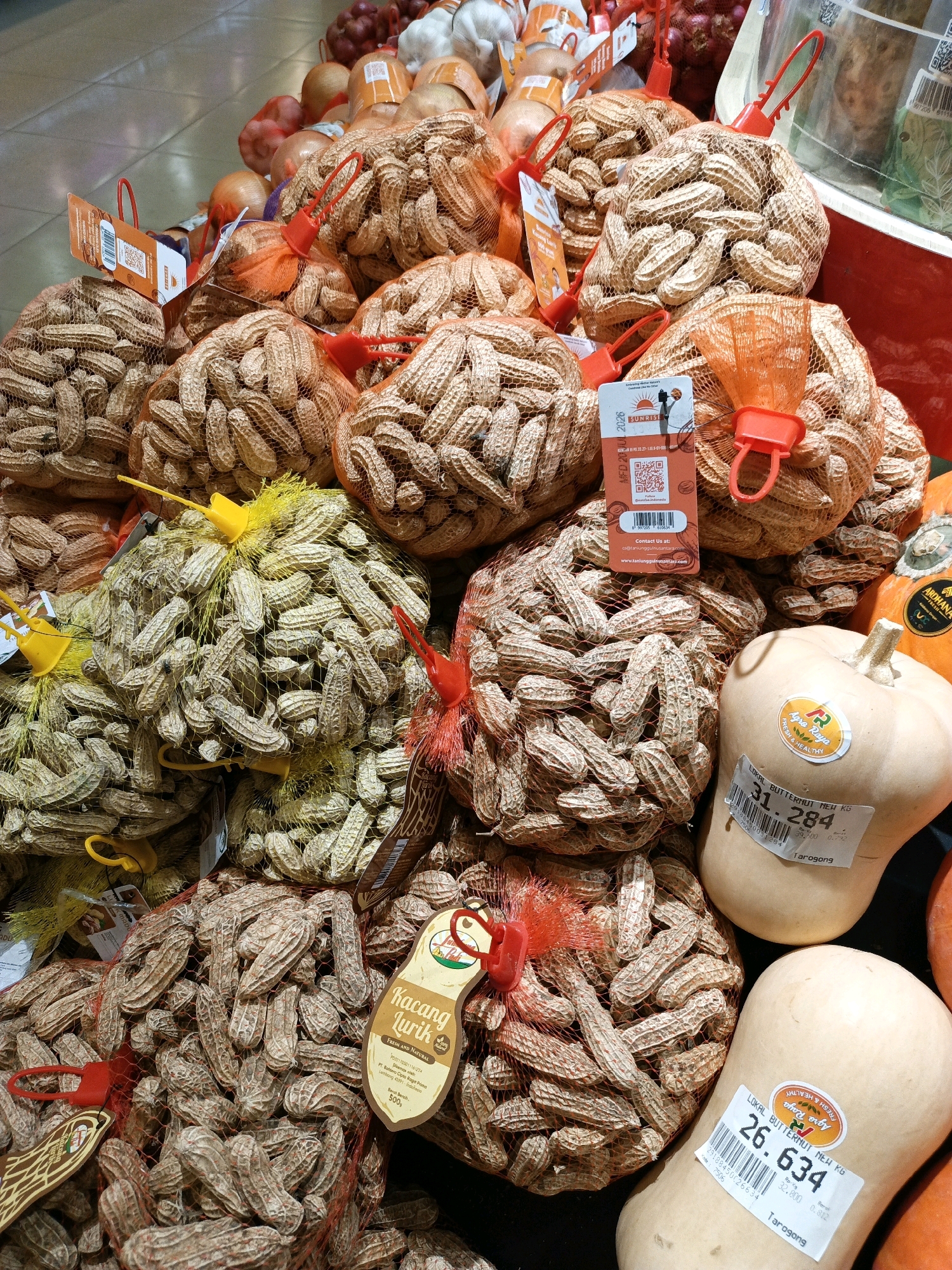 A close-up overhead shot captures a display of bulk produce, predominantly peanuts in mesh bags, alongside several butternut squashes. The peanuts are contained in various colored net bags – predominantly orange, but also yellow and red – with plastic closures and handles. Some bags have product tags with text and barcodes. In the foreground and mid-ground, multiple bags of peanuts are piled high, creating a textured landscape of shells. To the right, two pale yellow butternut squashes are visible, each with a label displaying a barcode, price, and product information, including "LOKAL BUTTERNUT MEI KG." One squash has a sticker with the text "Agro Raya FRESH & HEALTHY." In the background, out-of-focus items like onions and possibly garlic are visible, suggesting this is a market or grocery store produce section. The lighting is bright, illuminating the scene evenly. The overall impression is one of abundance and fresh produce. This scene is consistent with a grocery store produce aisle in South Jakarta, Indonesia.